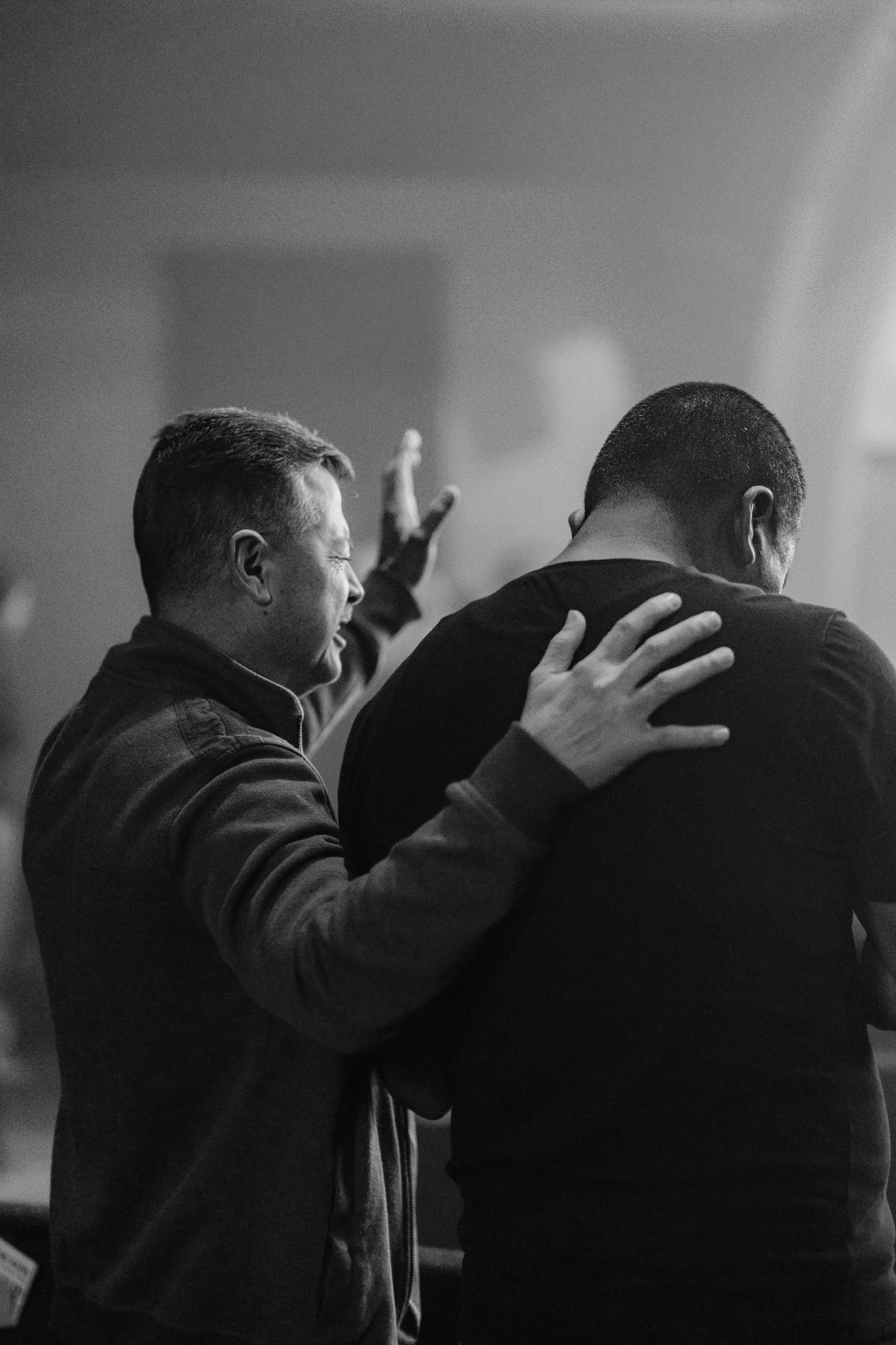 Two men in black jackets hugging and smiling warmly in a close embrace, one with his hand on the other's shoulder, in a dimly lit indoor setting.