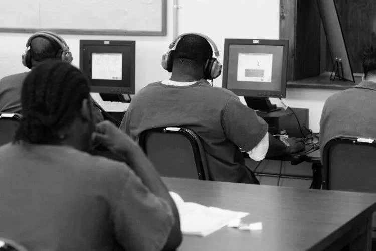 Students sitting in a classroom using desktop computers, wearing headphones, with some looking at their screens and others listening or observing.