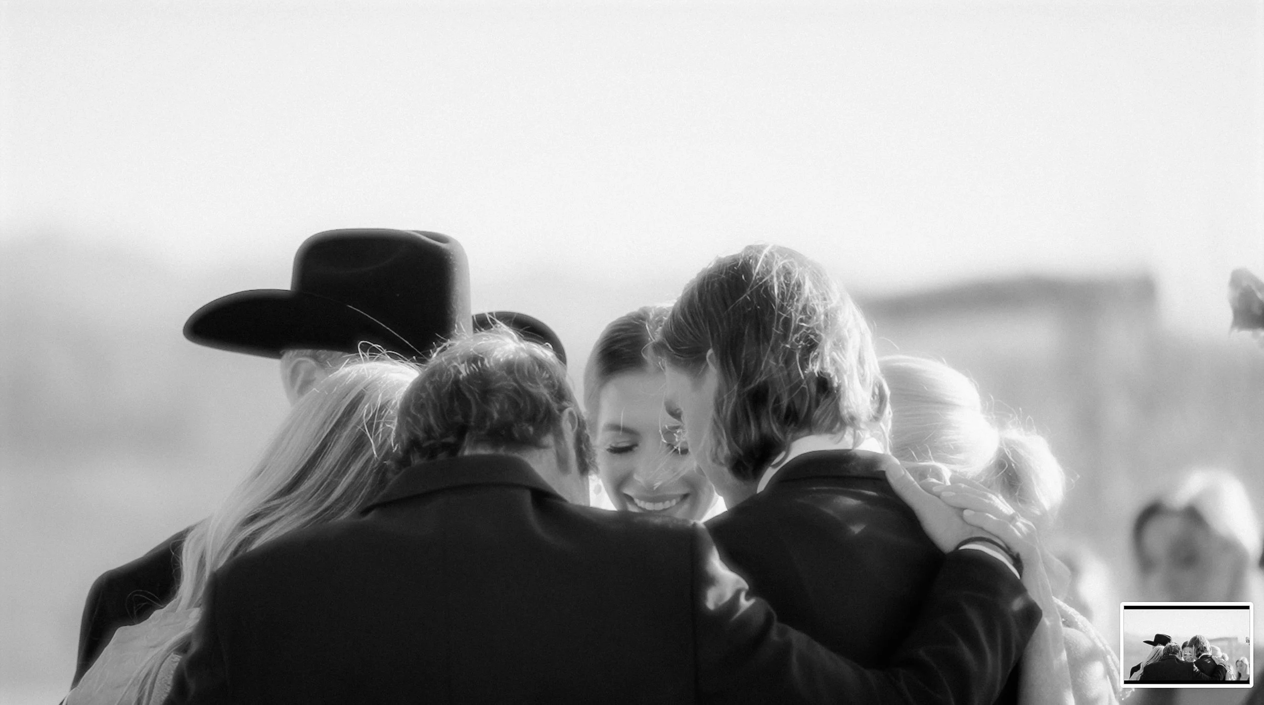 A black and white photo of a group of people embracing in a circle, with one person wearing a cowboy hat visible from the back.