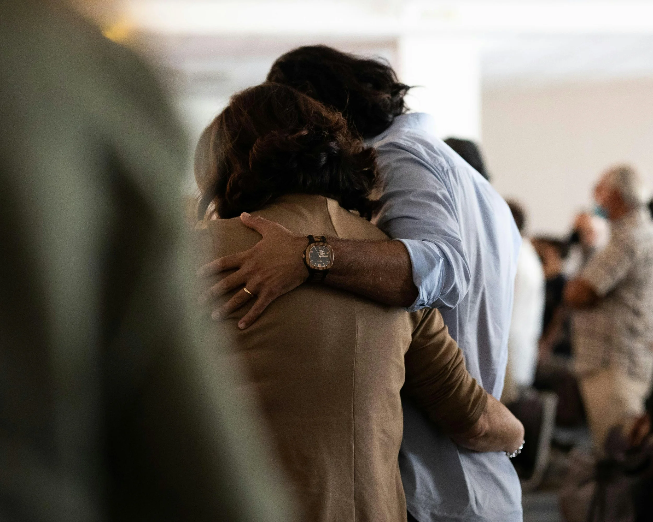 A close-up of two people hugging, showing only their heads and arms, in a room with other people in the background. One person with dark hair, the other with lighter hair, both wearing professional or casual attire.