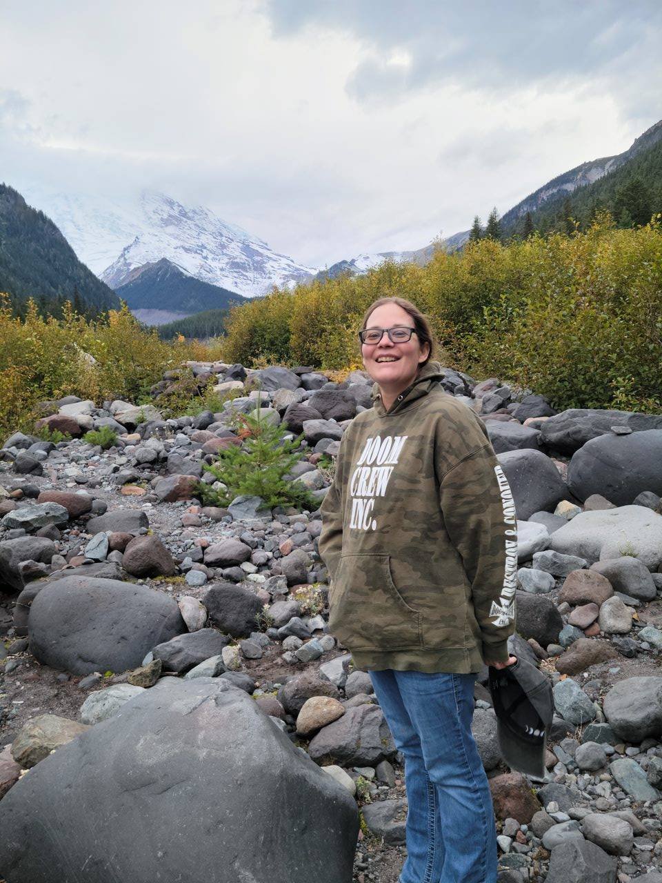 A woman smiling in a camouflage hoodie and jeans standing on rocky trail with mountains and snow-covered peaks in the background.