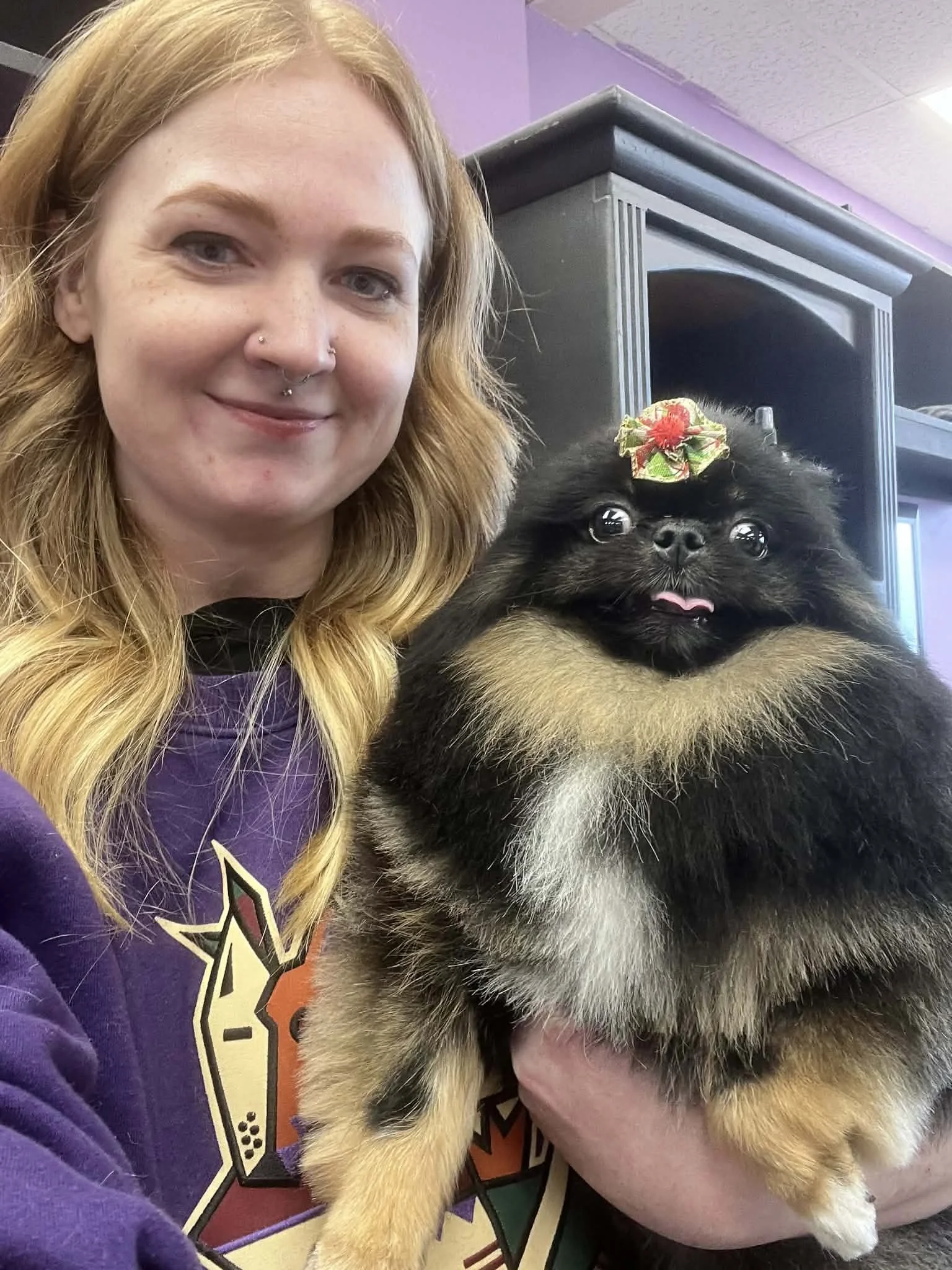 A woman with long red hair, light skin, and a nose piercing smiling next to a fluffy black and tan dog with a red and yellow bow on its head.