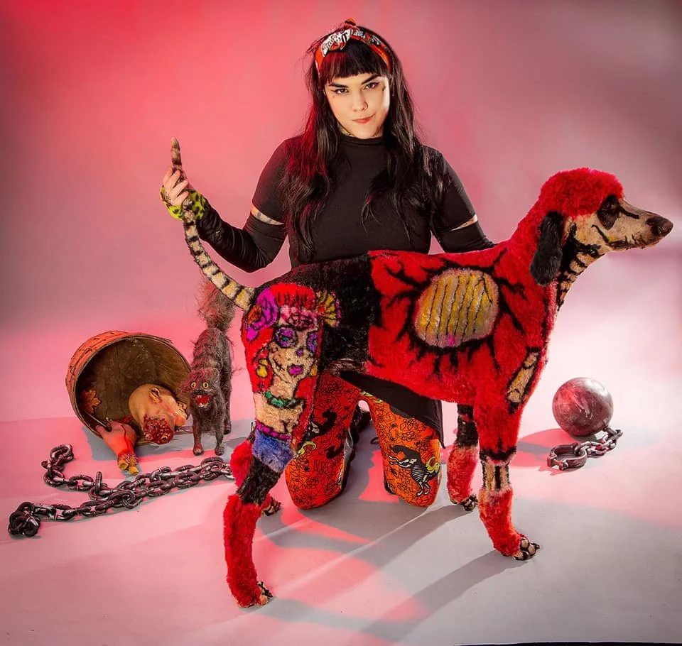 A woman with black hair and bangs wearing a red headband and black clothes, kneeling with her hand raised, surrounded by colorful art-covered dog and cat sculptures, a chained pig sculpture, a globe, and a small figurine, all set against a pink and purple background.