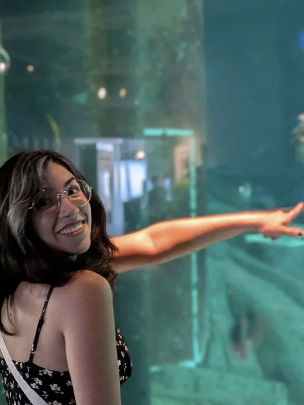 A woman with glasses smiling and pointing at an aquarium display.