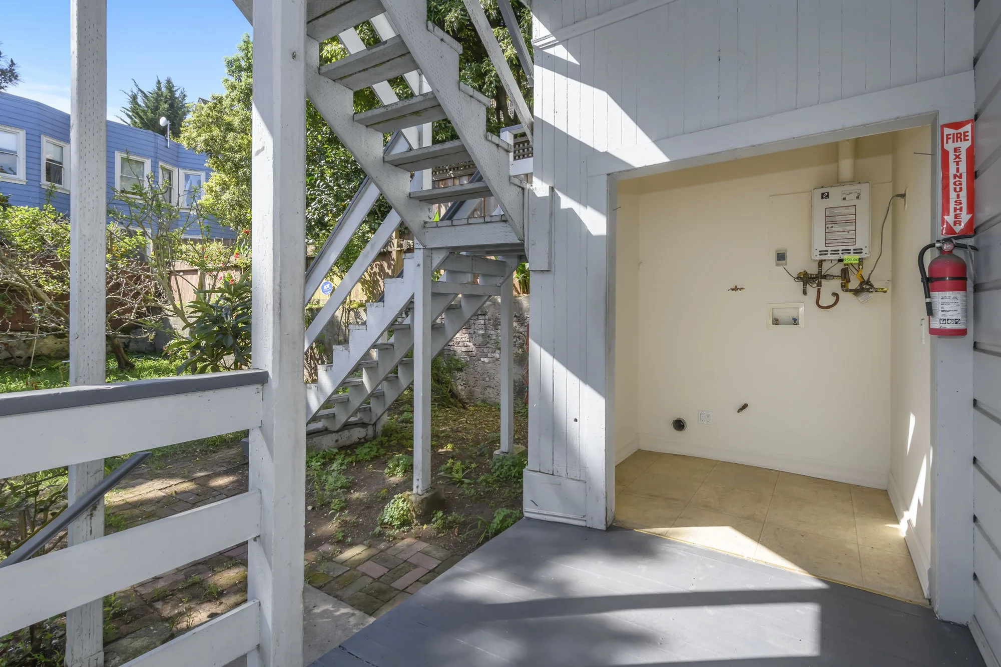 View of an outdoor staircase leading to an upper level, with a small, empty, enclosed area on the ground level featuring a water heater, fire extinguisher, and fire extinguisher sign, in a residential backyard with greenery and neighboring buildings 