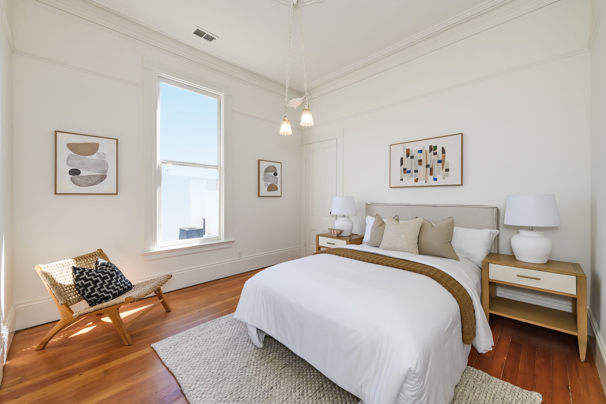 Bedroom with white walls, hardwood floors, a bed with white bedding and beige pillows, two wooden nightstands with white lamps, abstract artwork on the walls, a small woven bench with black and white pillows, and a beige rug.