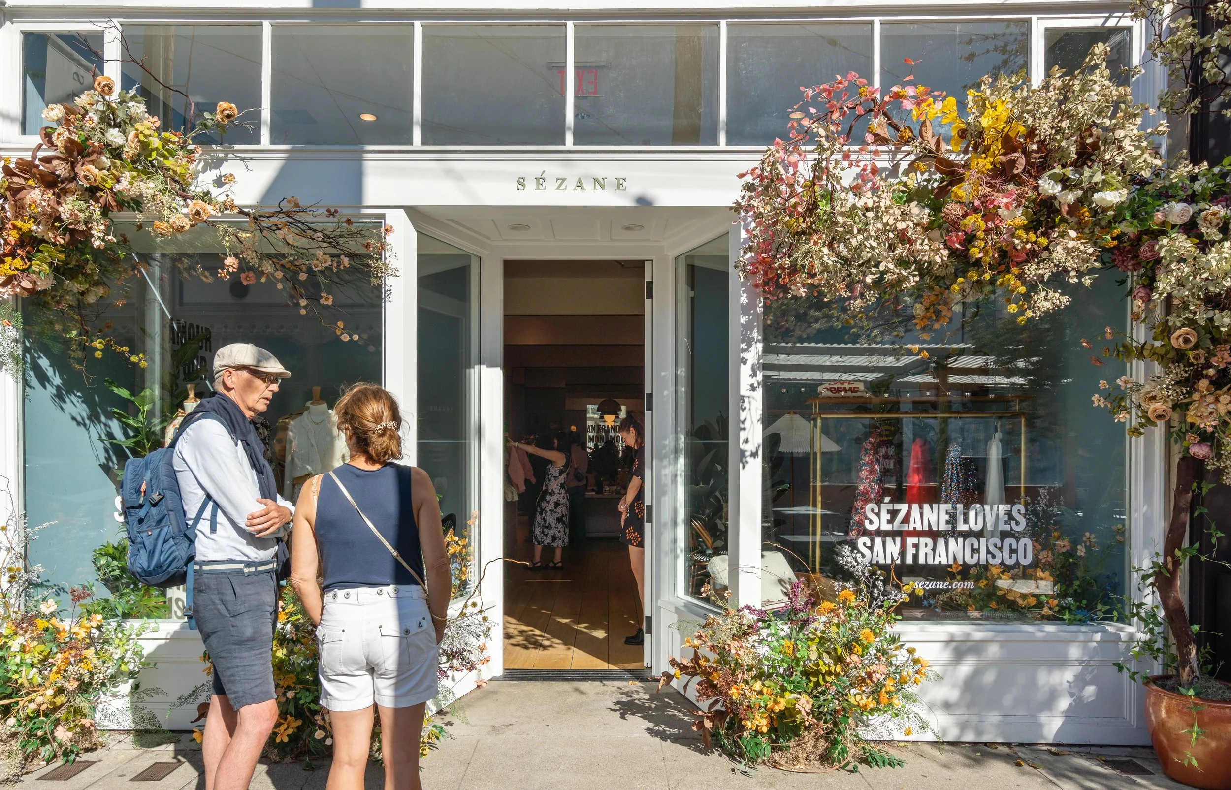 Two people standing outside the Sezane storefront, which has large glass windows decorated with floral arrangements. The sign reads 'Sezane Loves San Francisco' and the store interior shows shoppers and displays.