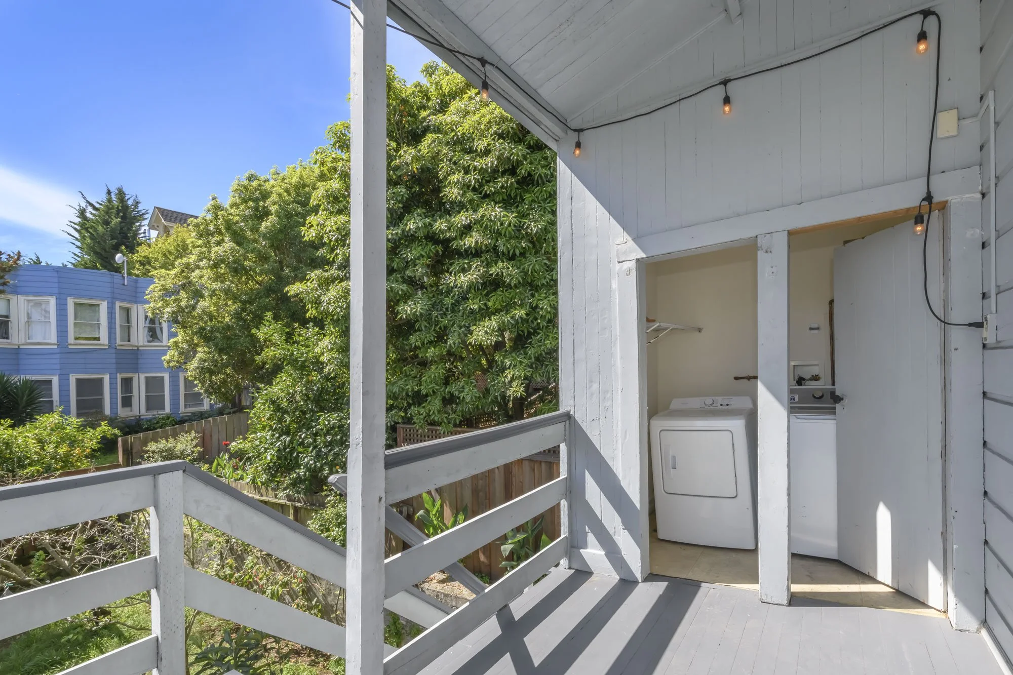 Balcony with white railing and laundry area, including a washing machine, in a house with siding, overlooking a green backyard with trees and a neighboring blue building