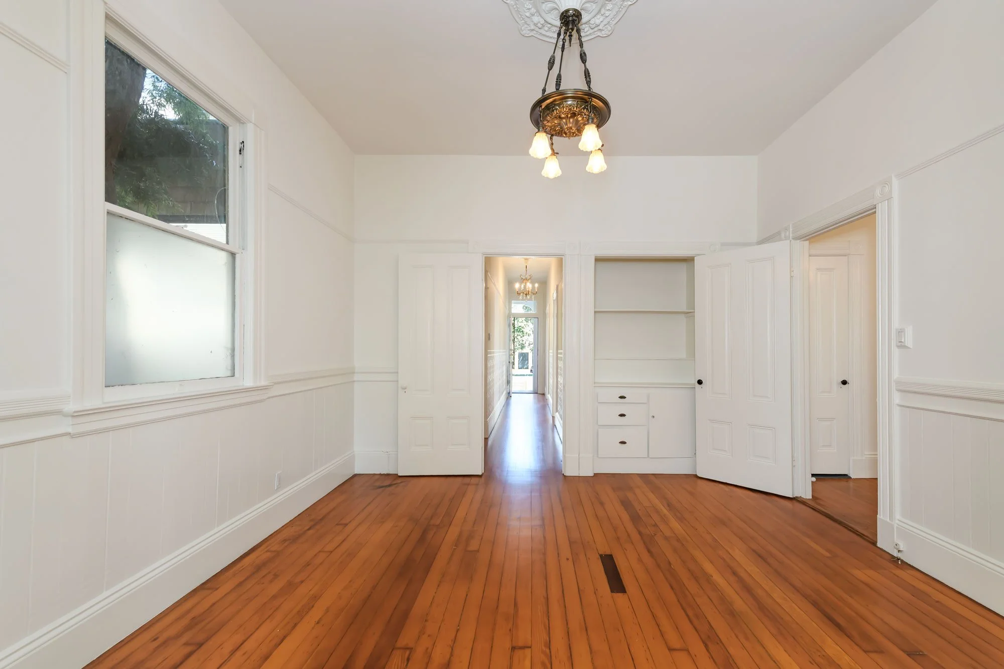 Empty room with hardwood floors and white walls, featuring a ceiling chandelier, a window on the left, and a doorway leading to a hallway with natural light.