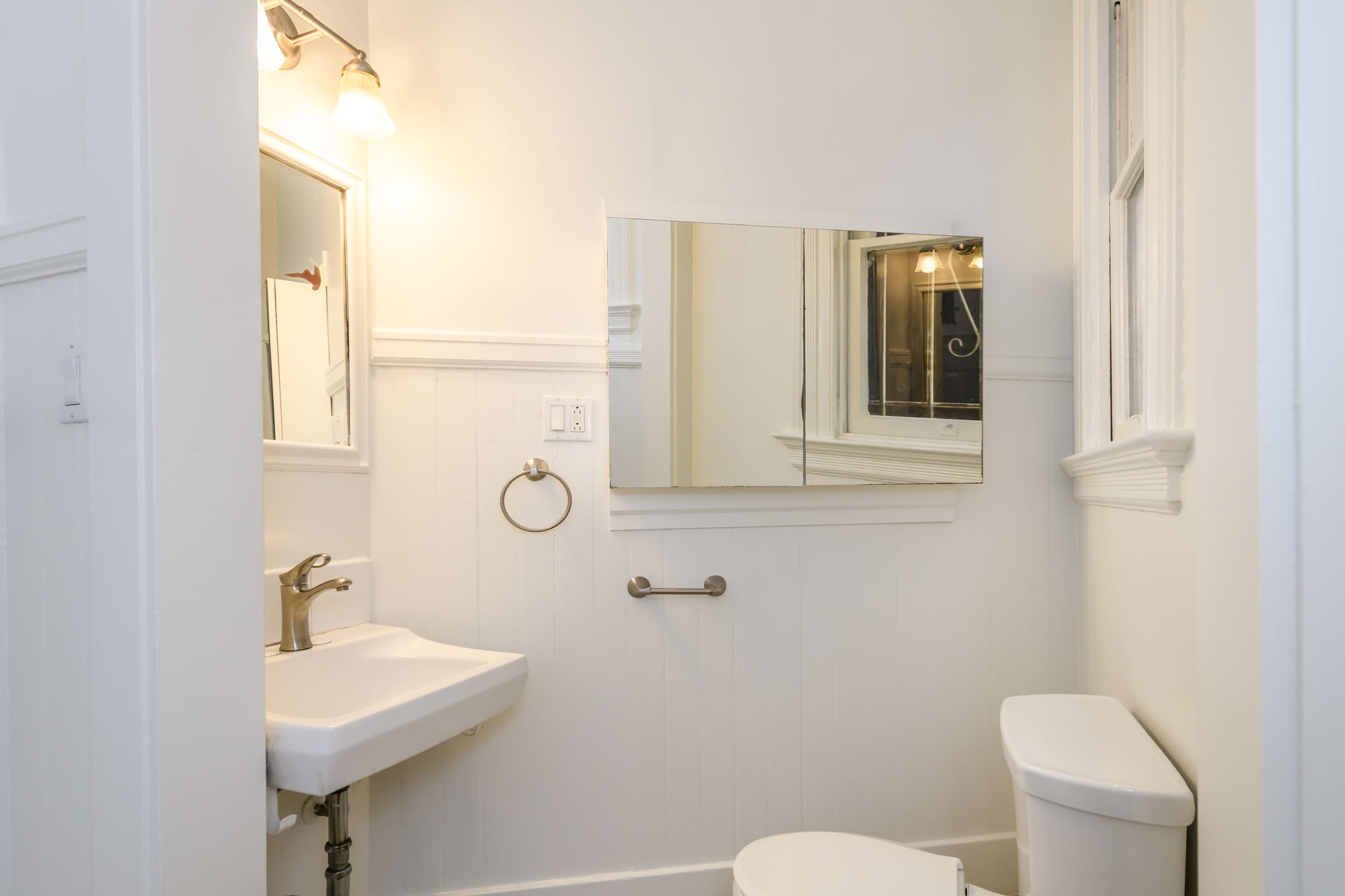 Small white bathroom with a wall-mounted sink, a mirrored cabinet, a toilet, and a window.