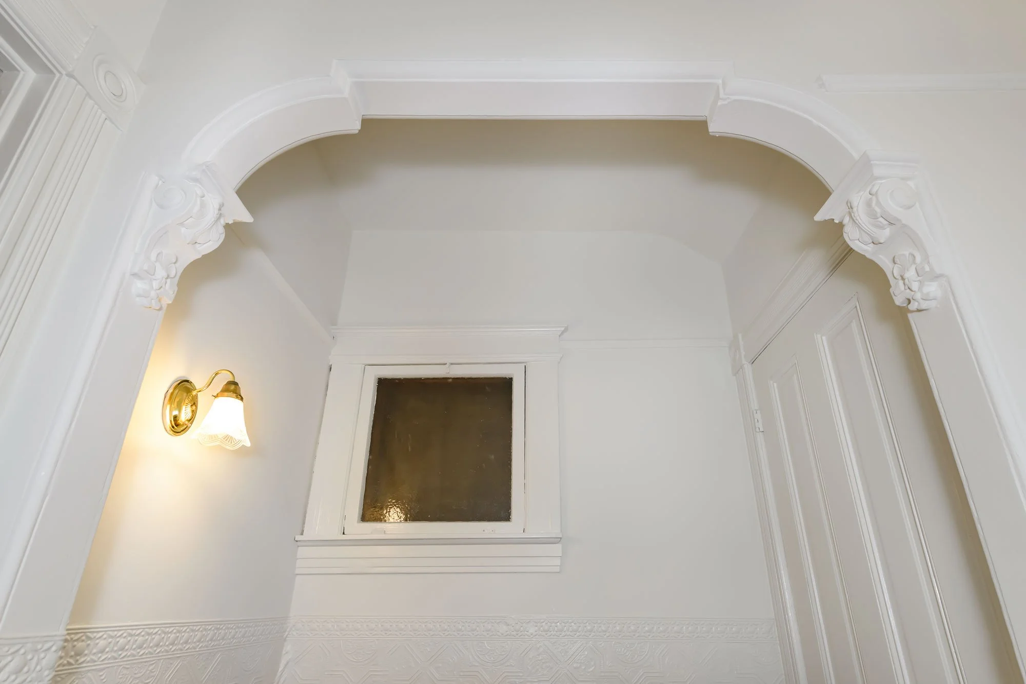 Interior view of a white room with ornate architectural molding and trim, a small window, a brass wall sconce with a frosted glass shade, and crown molding near the ceiling.