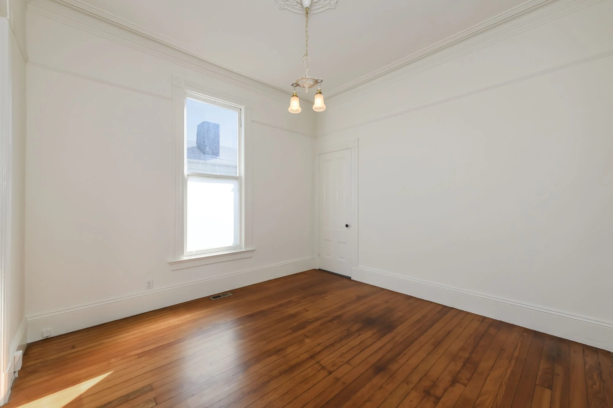 Empty room with white walls, hardwood floor, large window, and ceiling light fixture.