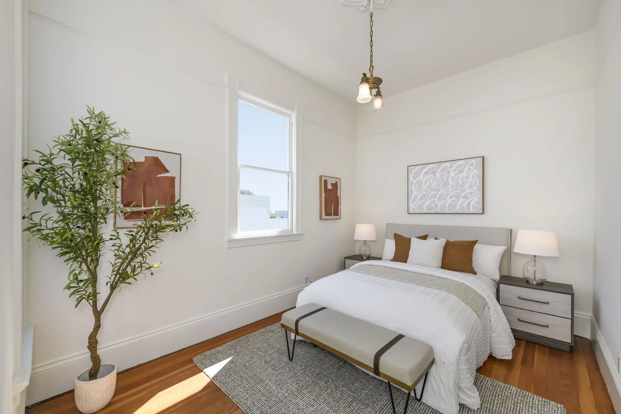 A neatly made bed with white and brown pillows, two nightstands with lamps, a plant in a pot, and a bench at the foot of the bed in a bright bedroom with white walls and wooden floors.