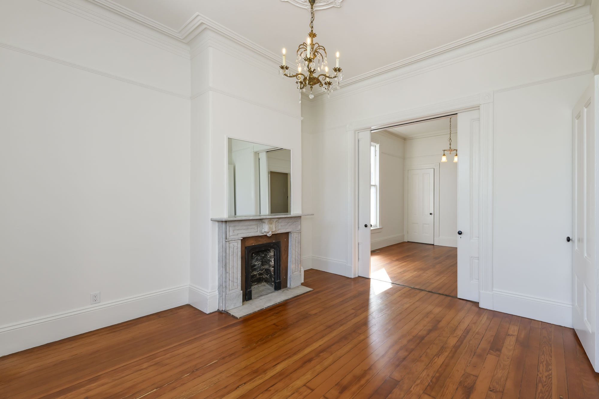 Empty room with hardwood floors, a white fireplace with a mirror above it, and a chandelier hanging from the ceiling.