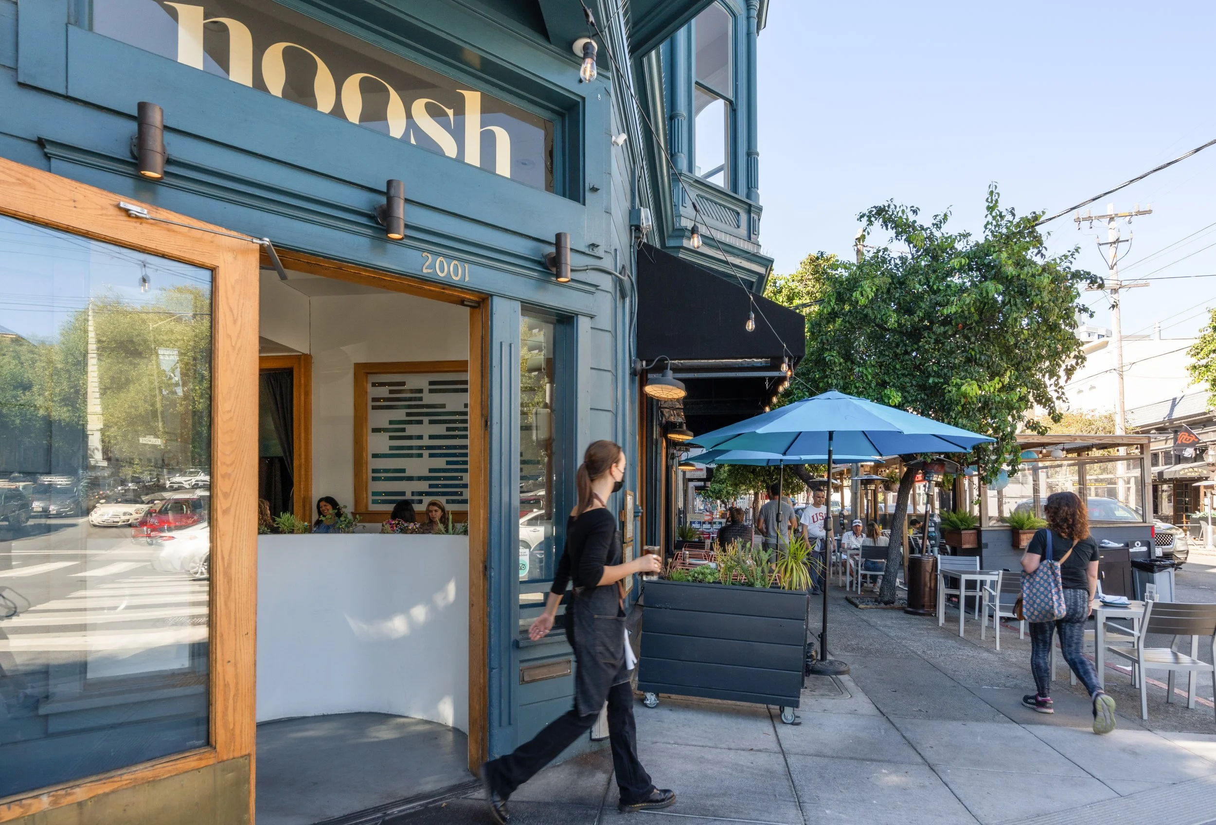 People walking outside a coffee shop with outdoor seating, blue building exterior, and a large blue umbrella on the sidewalk.