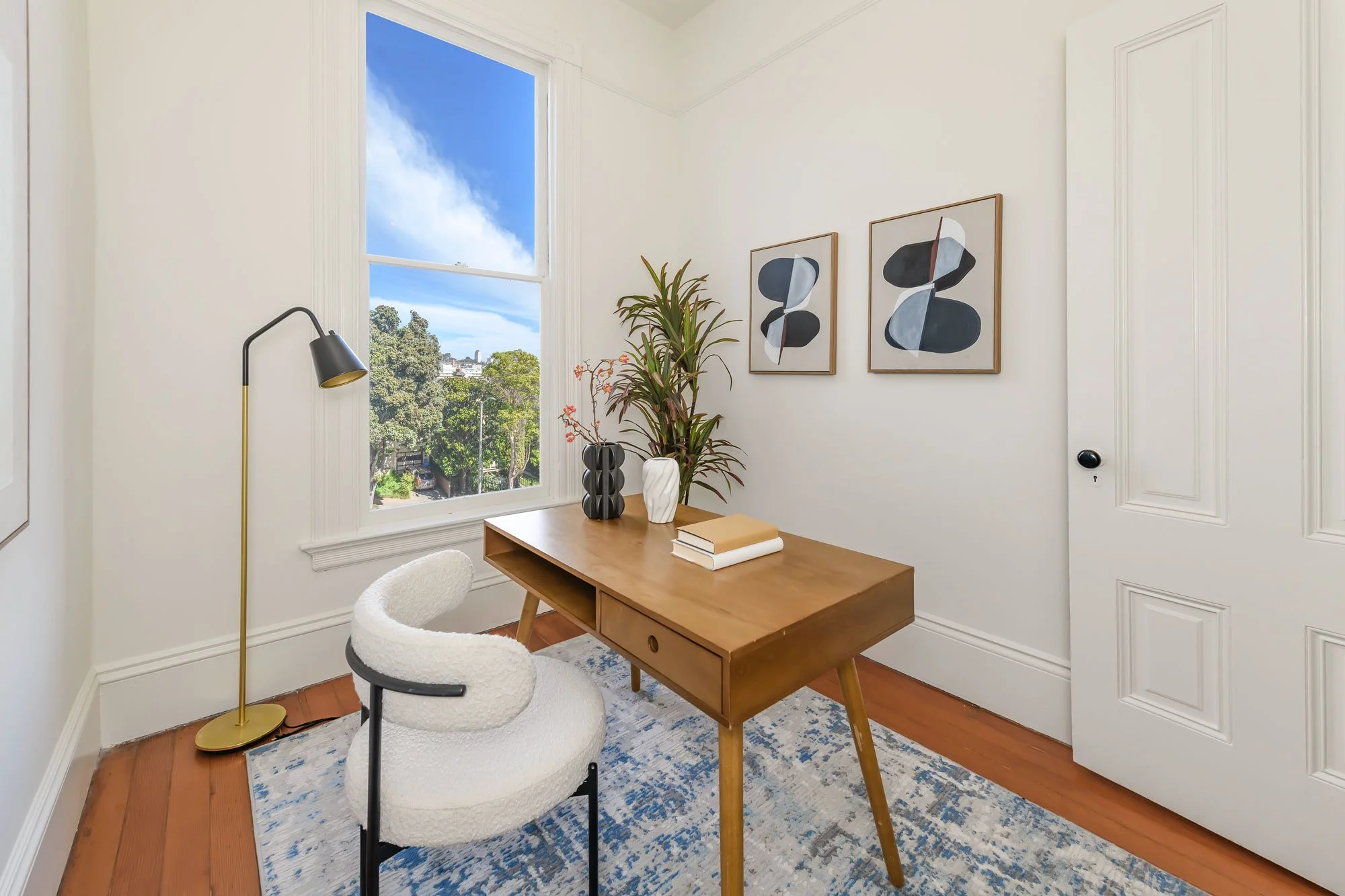 A home office with a wooden desk, a plush white chair, a tall window with a view of trees and blue sky, three framed abstract art pieces on the wall, and a tall plant in a white vase on the desk.