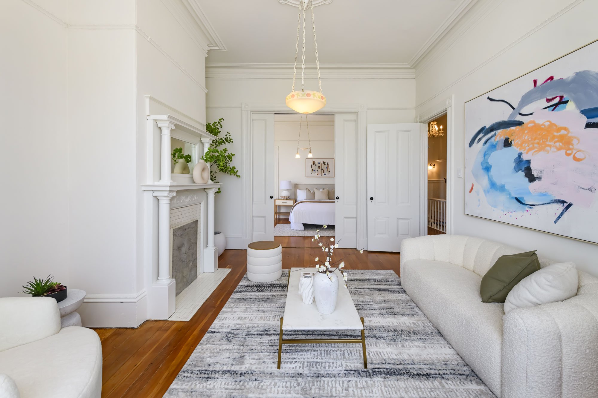Living room with white walls, hardwood floors, a white sofa with pillows, a marble-top coffee table with vases, colorful abstract painting on the wall, and view into a bedroom with a bed and nightstand.