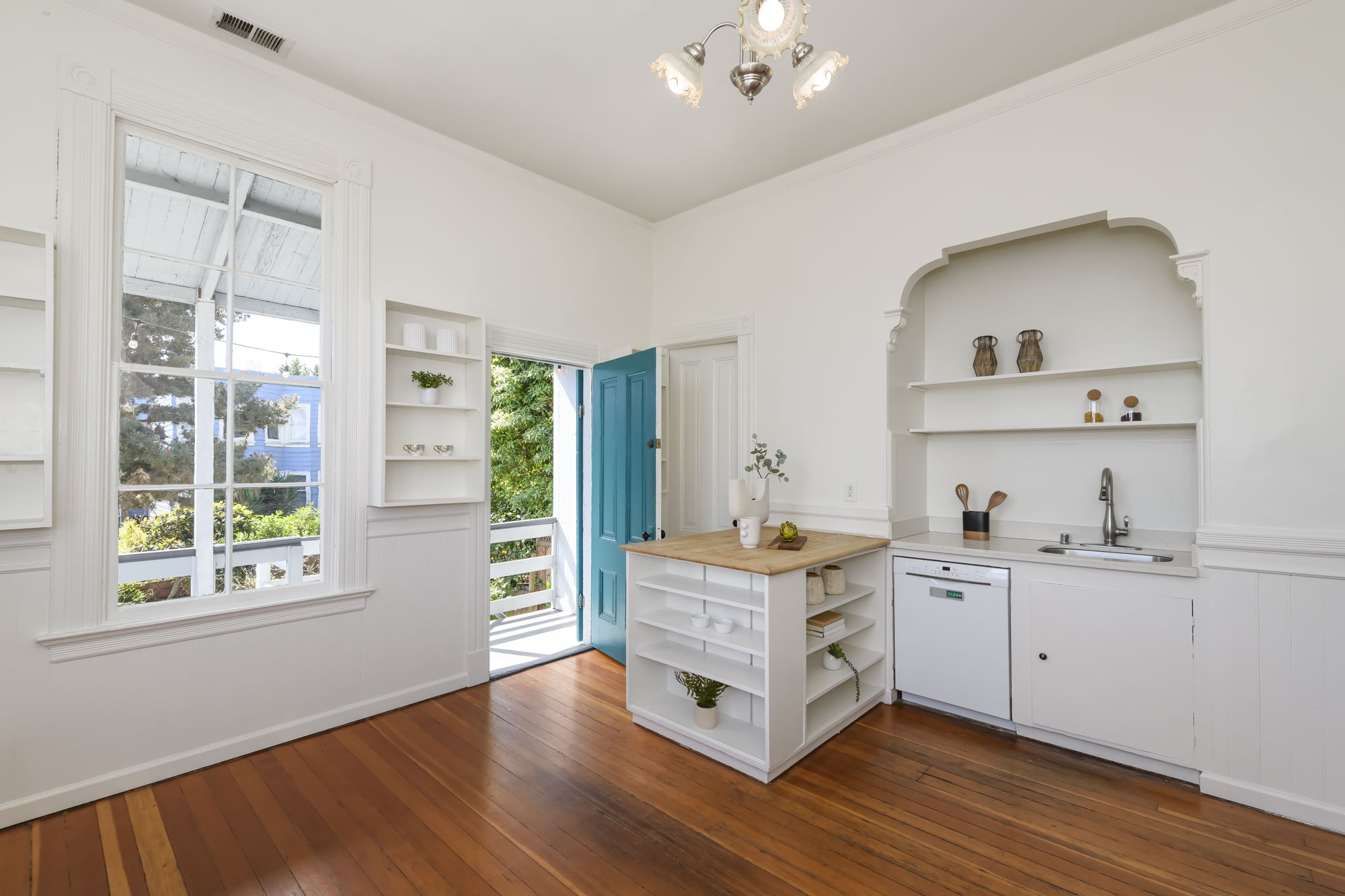 A bright, minimal kitchen with hardwood floors, white walls, and a blue front door. There are large windows and open shelving with decorative items, a small kitchen island, and a small sink area.