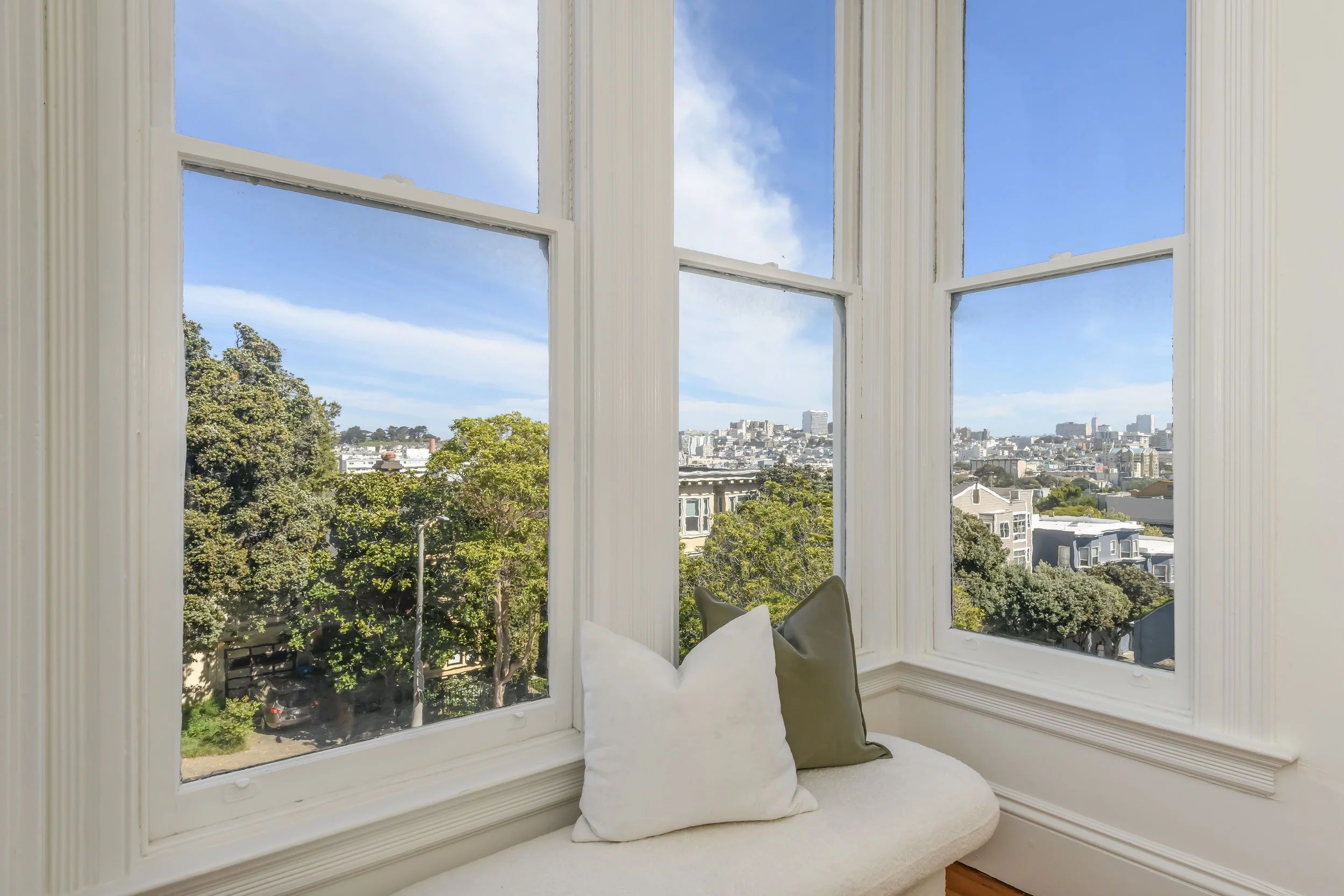 View of a cityscape through large white-framed windows, with a window seat and two pillows in the foreground.
