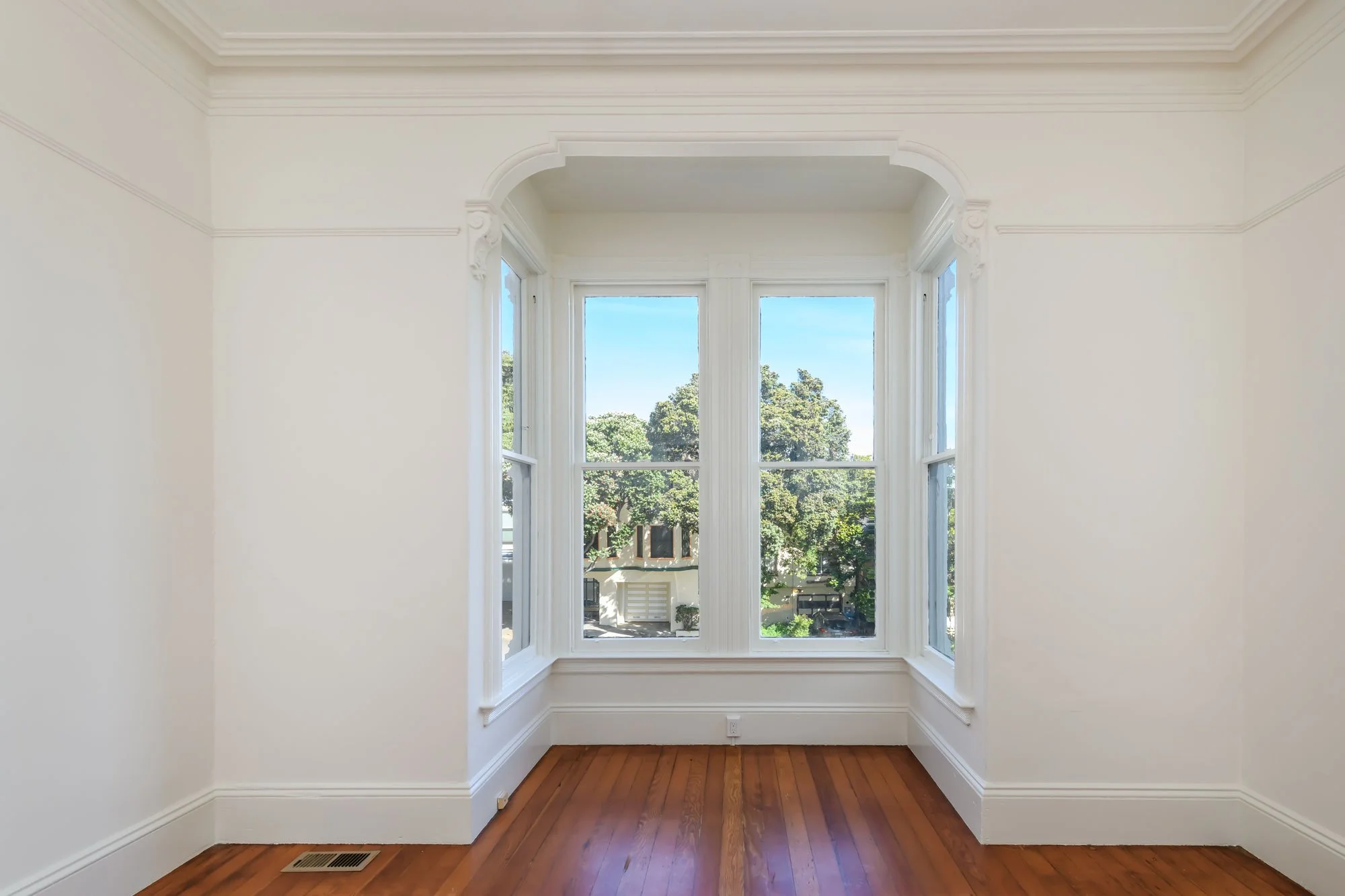 Empty room with white walls, wooden floor, large window with three panels showing trees and a building outside.