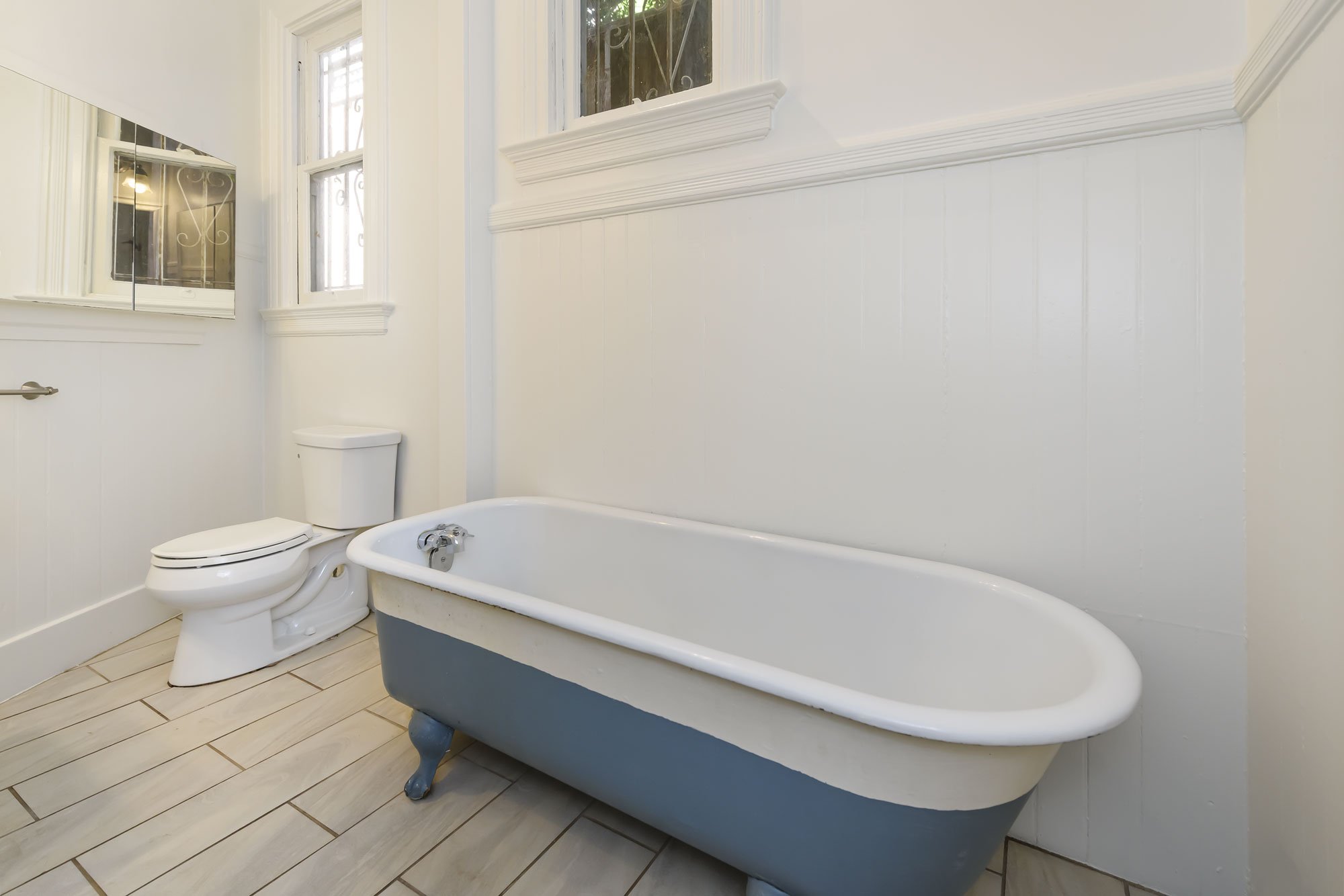Bathroom with a white bathtub, a toilet, a window, a mirror, and beige tile flooring.