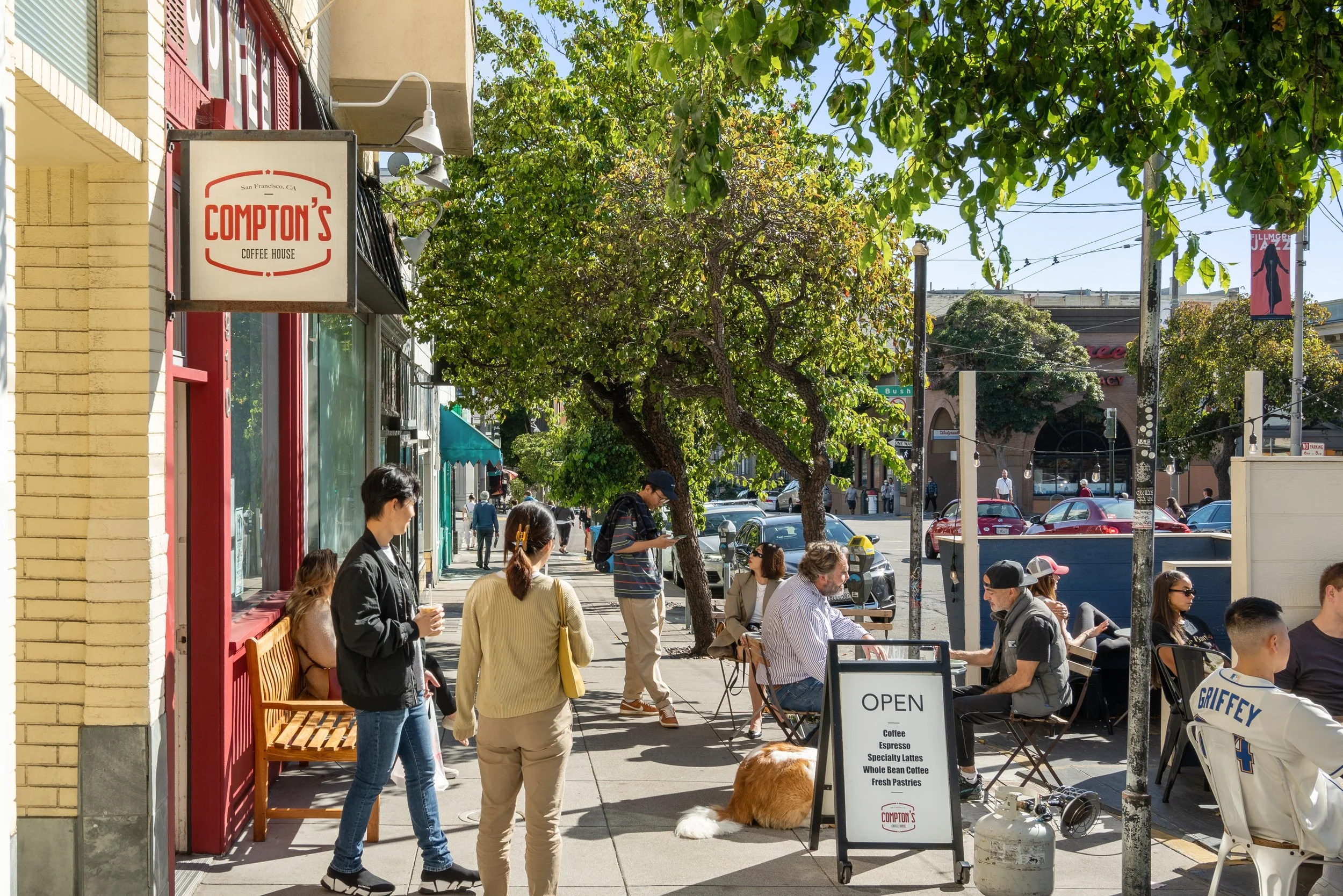 People sitting and standing outside a coffee shop with a sign that reads 'Open' and another sign for 'Compton's Coffee House'. Some are chatting, one person is reading a menu, and a dog is lying on the sidewalk.