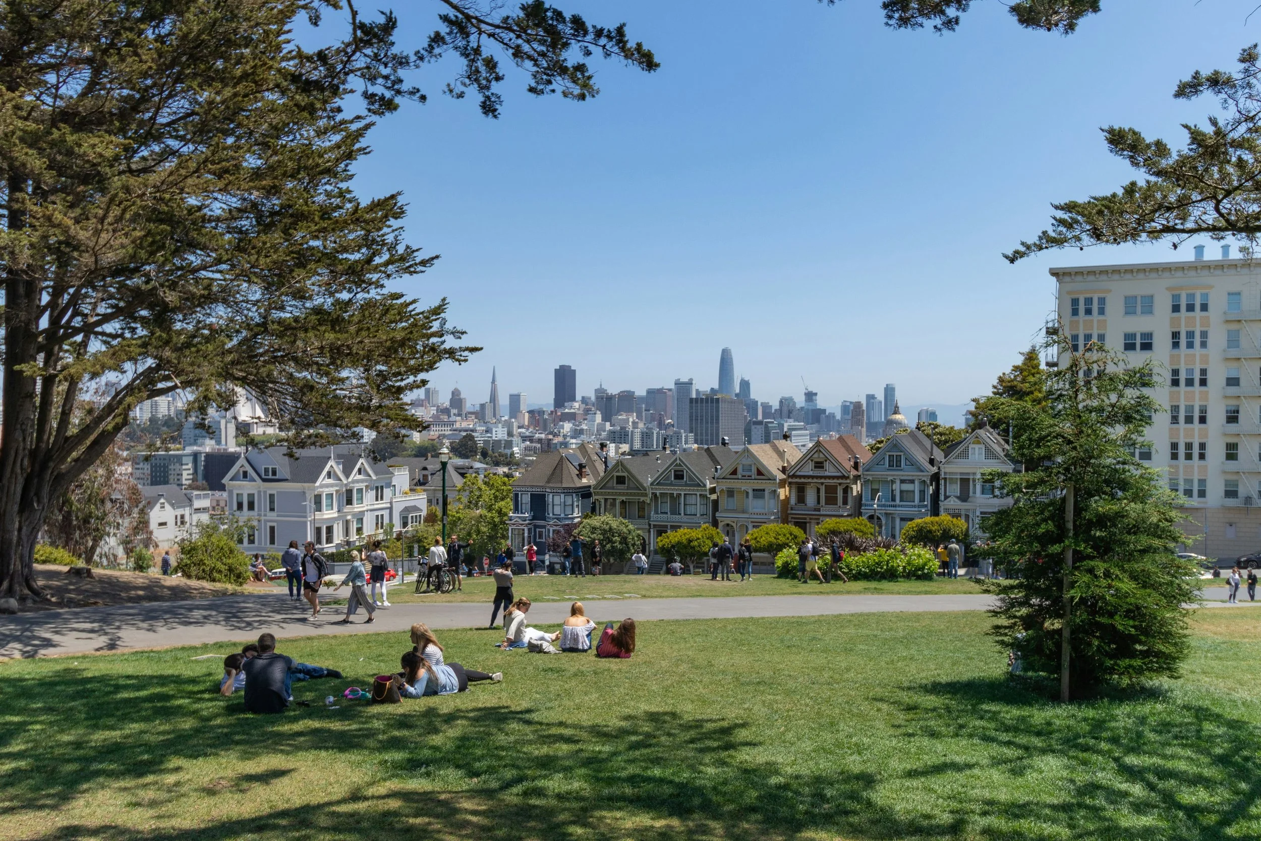 People relaxing on the grass and walking in a park with a city skyline in the background, including tall buildings and lush green trees.