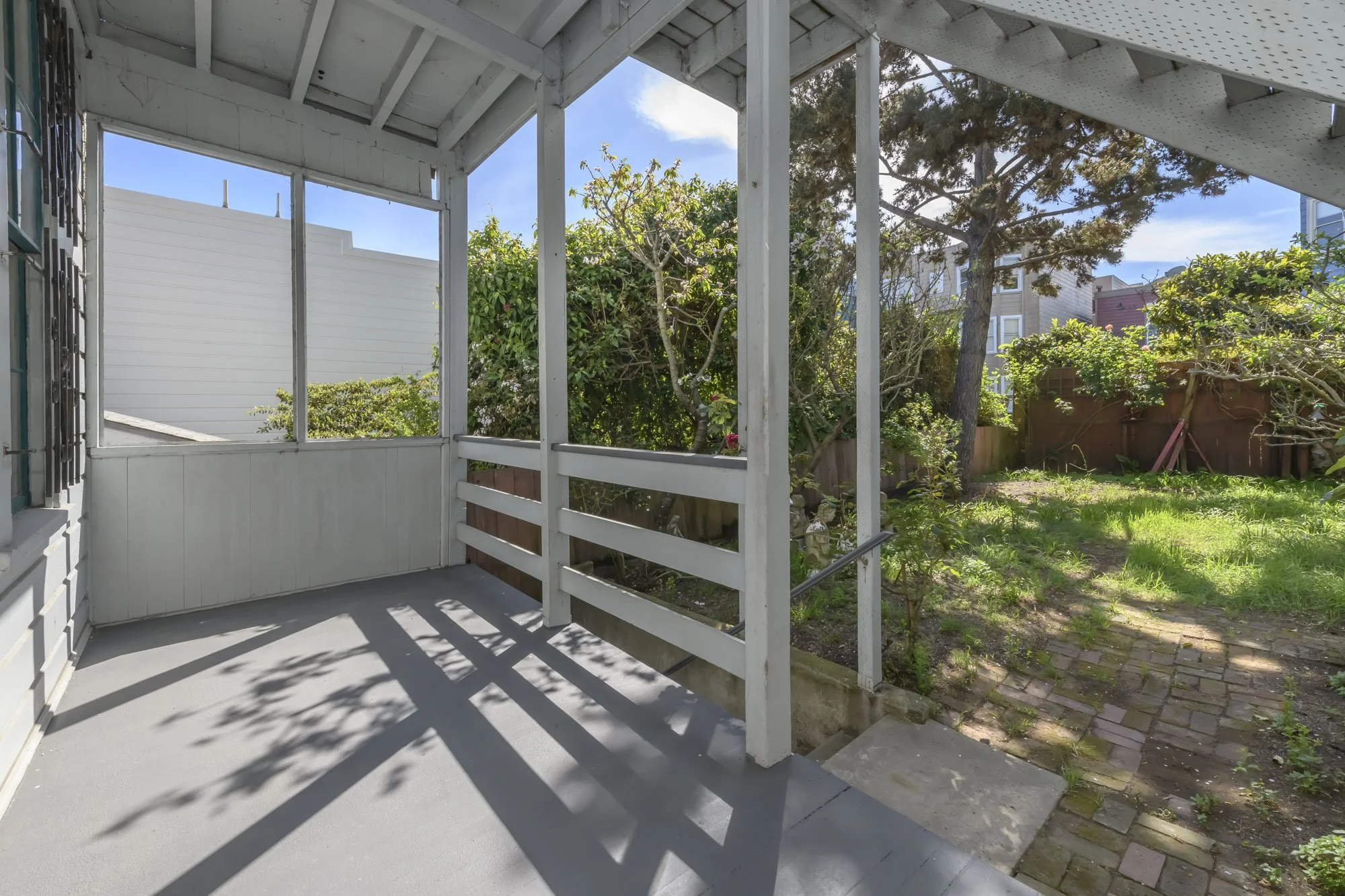 View from a porch looking out onto a garden with trees, grass, and a brick pathway under a partly cloudy sky.