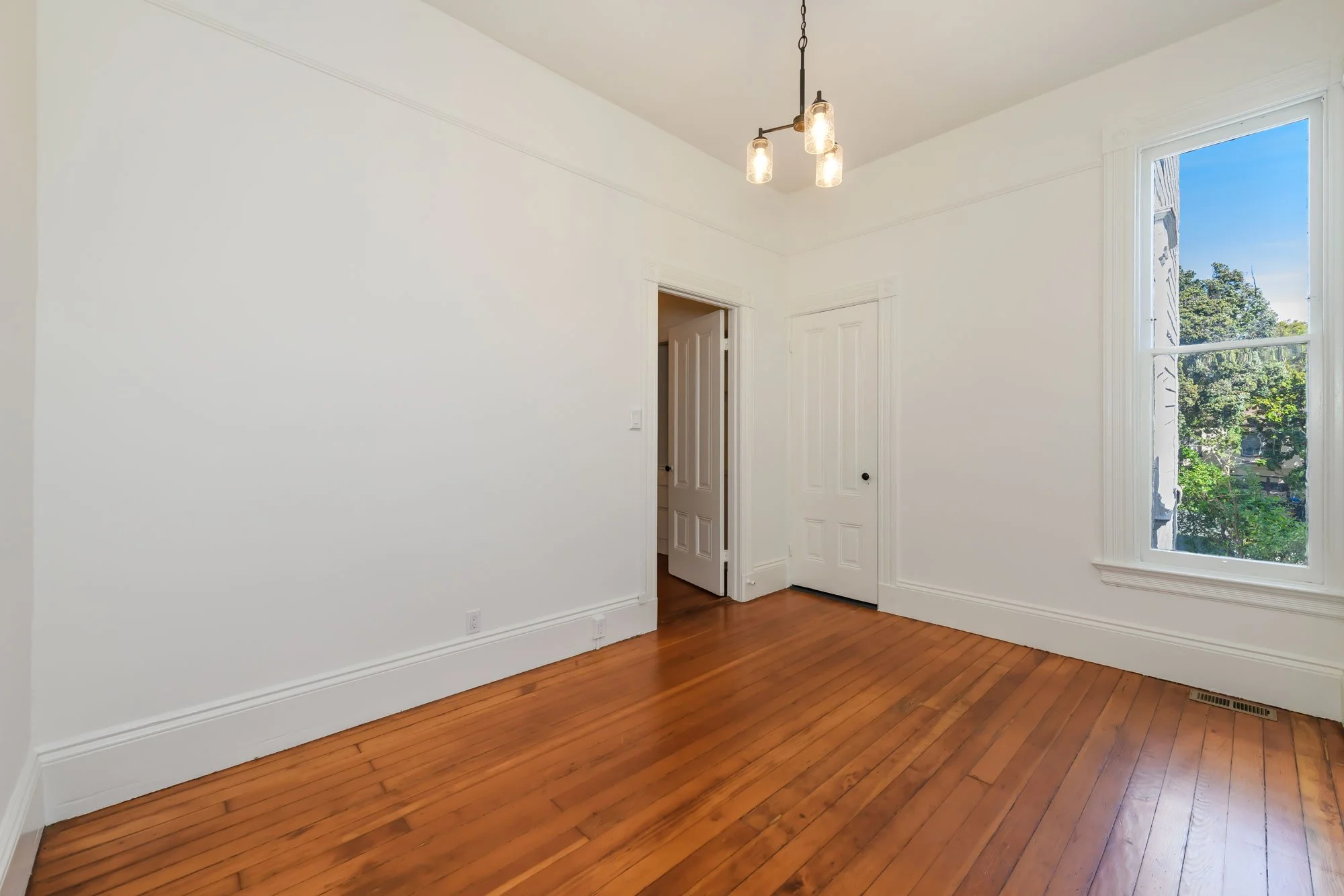 Empty room with white walls, hardwood floor, large window showing outdoor trees, chandelier lighting, and a door leading to another room.