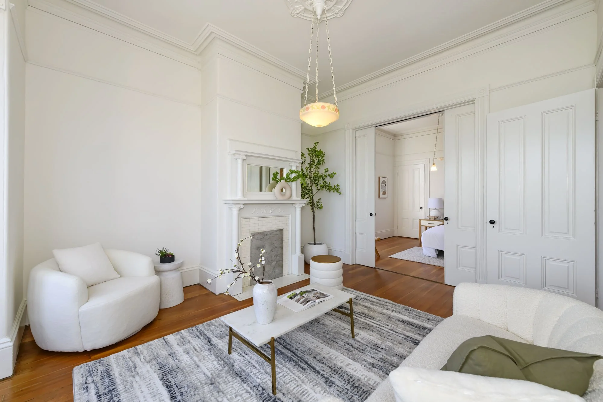Bright living room with white walls, a white sofa, and a marble fireplace. Decor includes vases, a small plant, and a rug. An archway leads to a bedroom with a bed and nightstand.