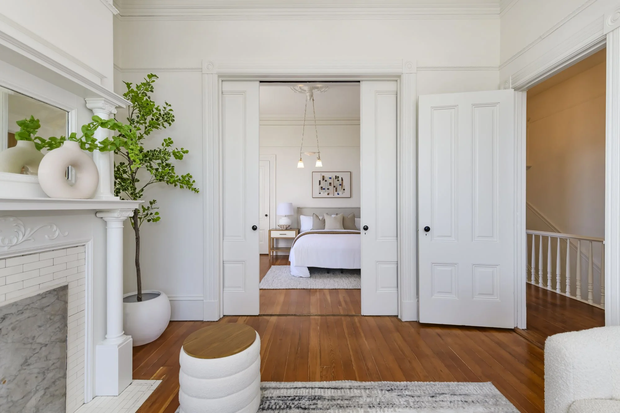 Interior view of a white bedroom with wooden flooring, seen through open sliding doors from a living room. The bedroom has a bed with decorative pillows, a nightstand with a lamp, and wall art. The living room features a fireplace with vases and a la