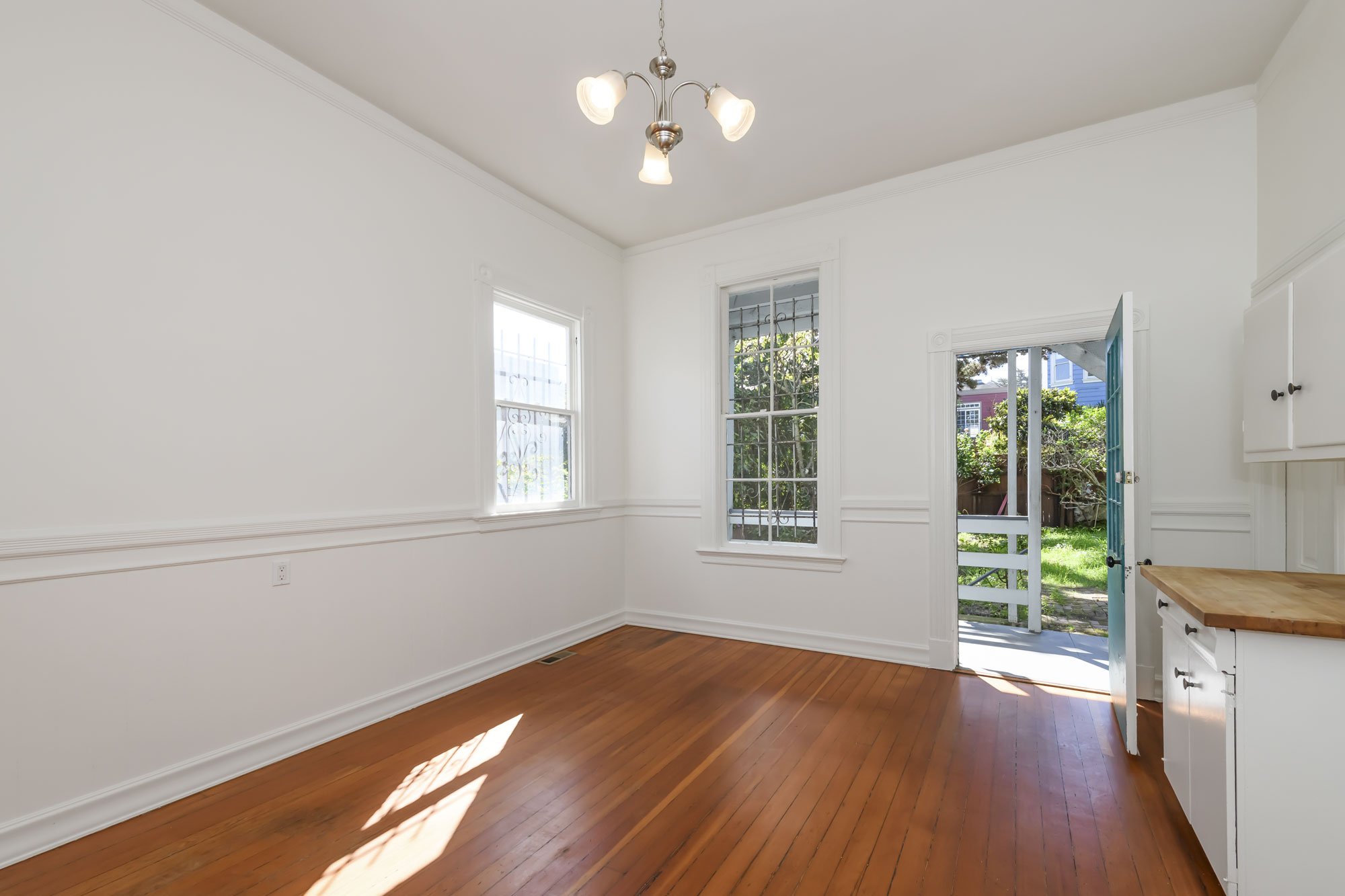 Empty room with wooden floors, white walls, two windows, a ceiling light fixture, and a door leading outside to a green yard.