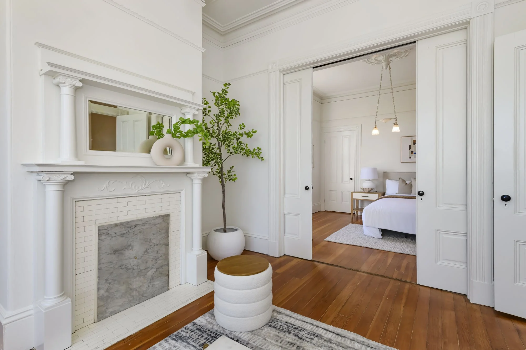 A bright and airy living room with white walls and trim, a fireplace with a white mantel and marble interior, a potted tree, a small round ottoman with a wooden top, and a mirror above the fireplace. An open doorway reveals a bedroom with a bed, nigh