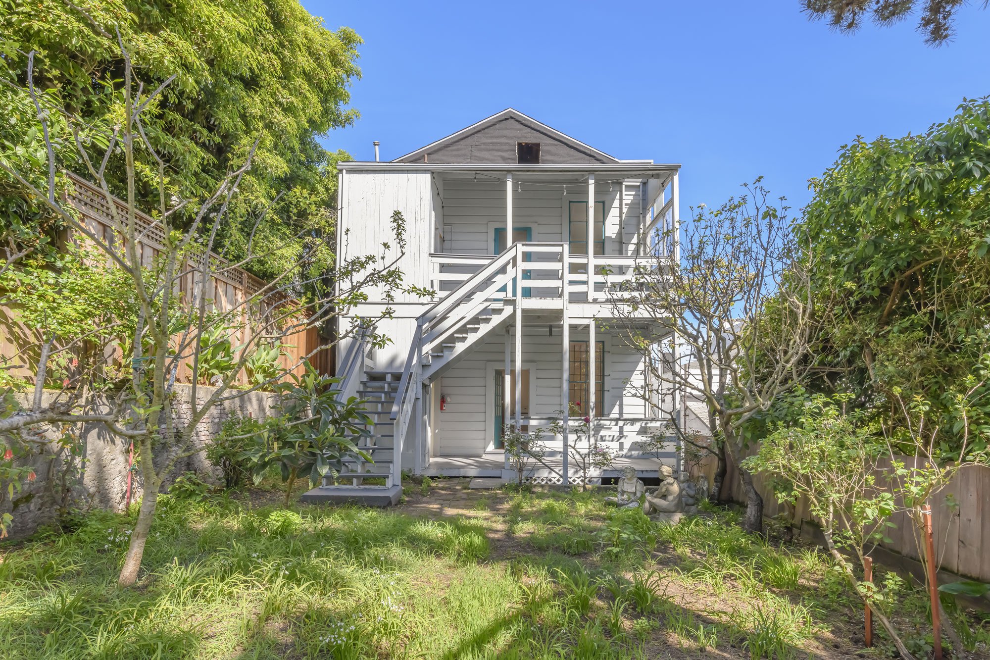 White two-story house with stairs leading to the upper porch, surrounded by green trees and plants on a sunny day.