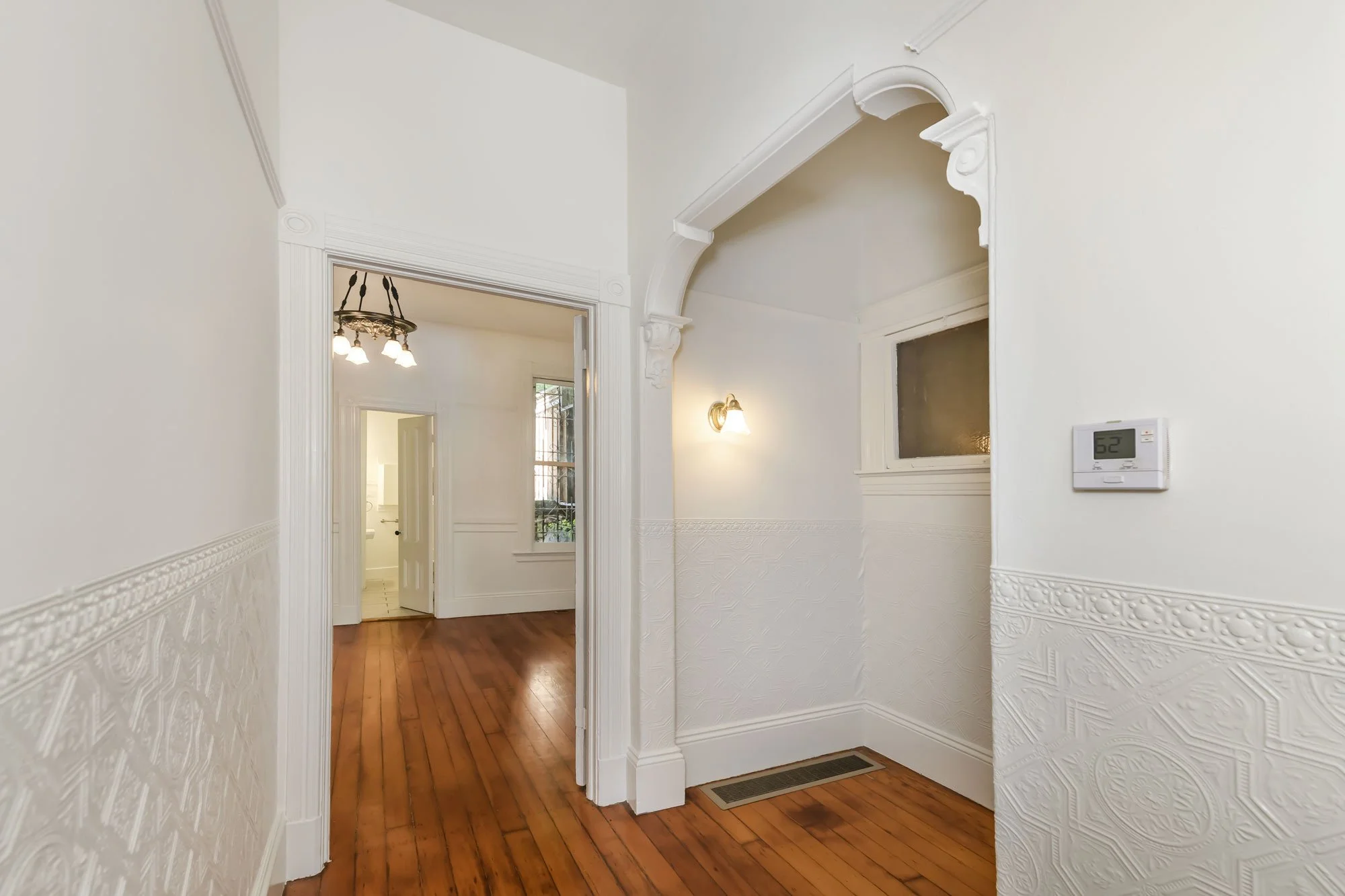 Interior view of a house hallway with white walls, wooden floor, a thermostat on the wall, decorative molding, and a doorway leading to another room.