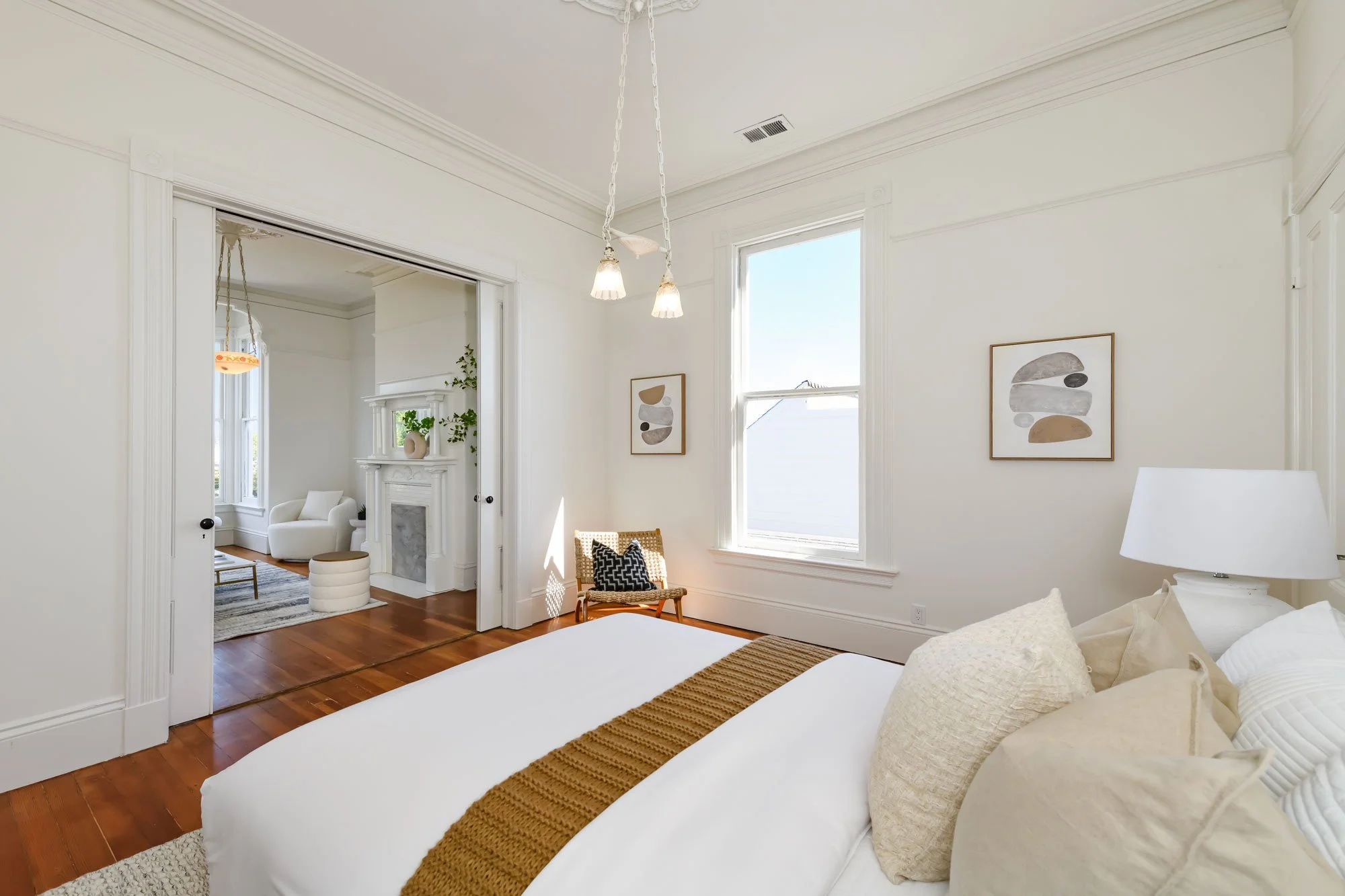 A bright, minimalist bedroom with white walls, a white bed with neutral pillows and a brown throw blanket, a white bedside lamp, and artwork on the wall. There is a wooden chair with a black and white cushion near the window, and an adjacent room wit