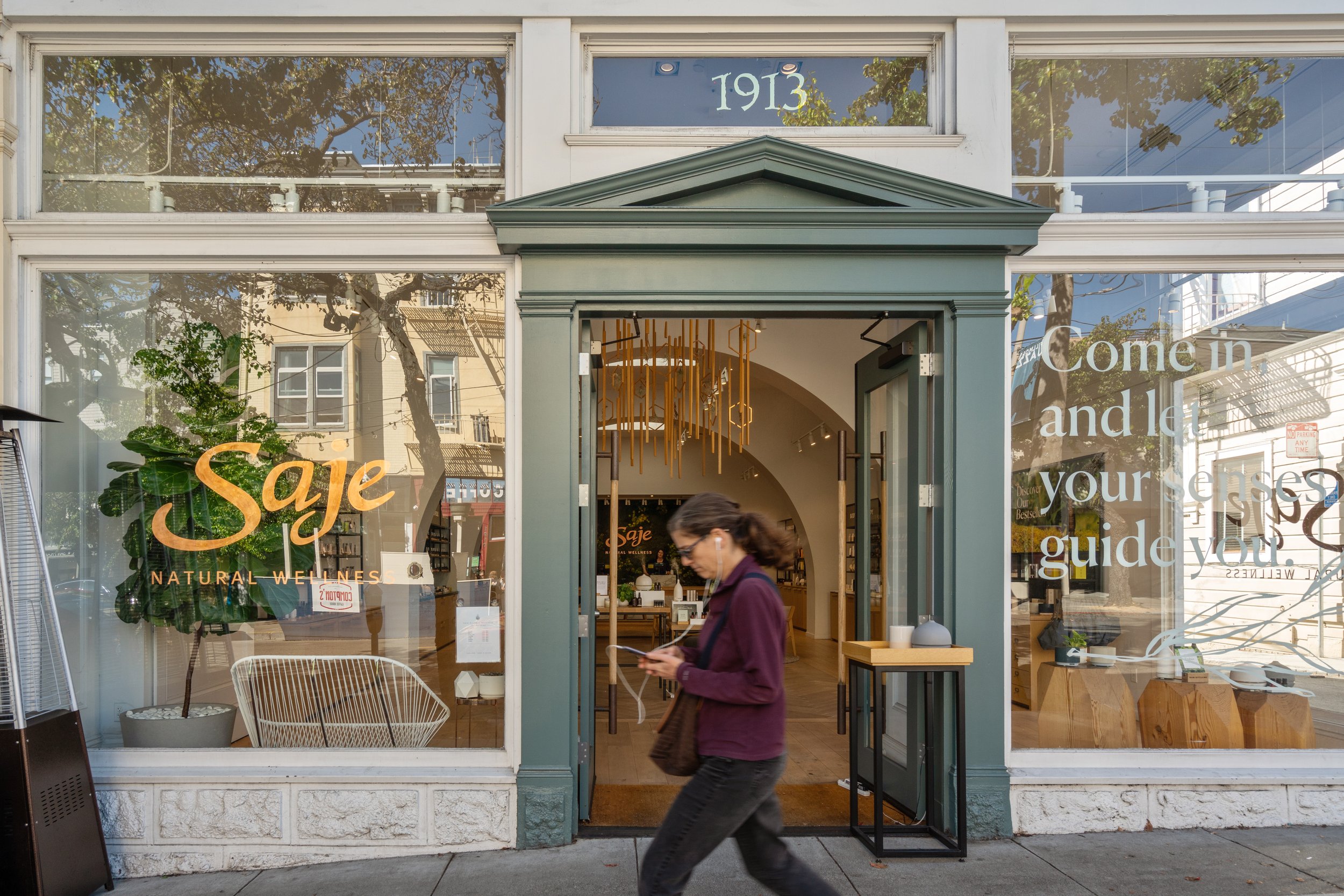 A woman walking past the storefront of Saje Natural Wellness with large glass windows and an open door, displaying interior decor and products, with the year 1913 visible above the entrance.