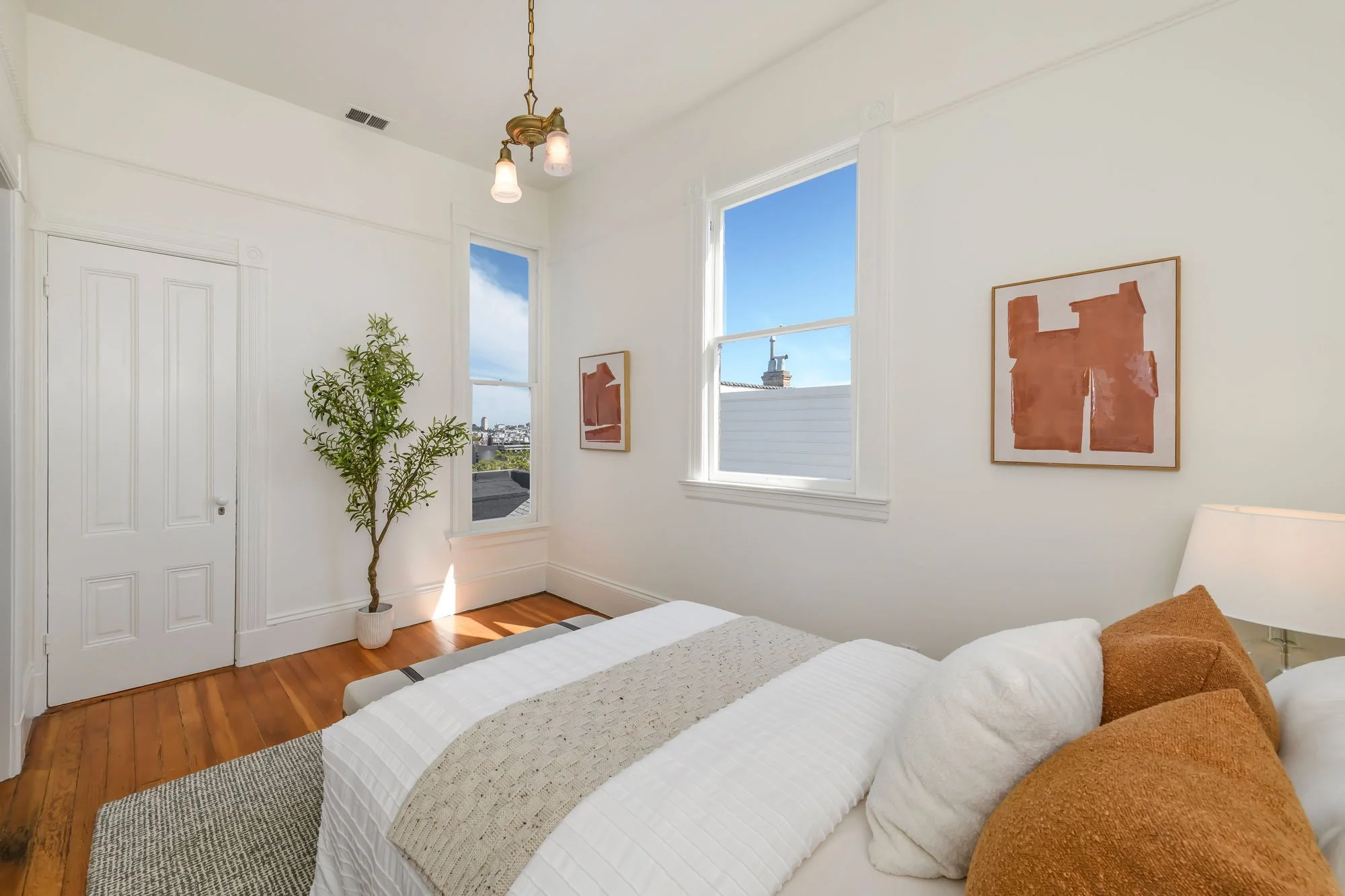 A bedroom with two white windows, a white bed with white and beige bedding, brown pillows, a white lamp, an abstract orange painting on the wall, a potted plant, wooden floors, and a ceiling light with three bulbs.