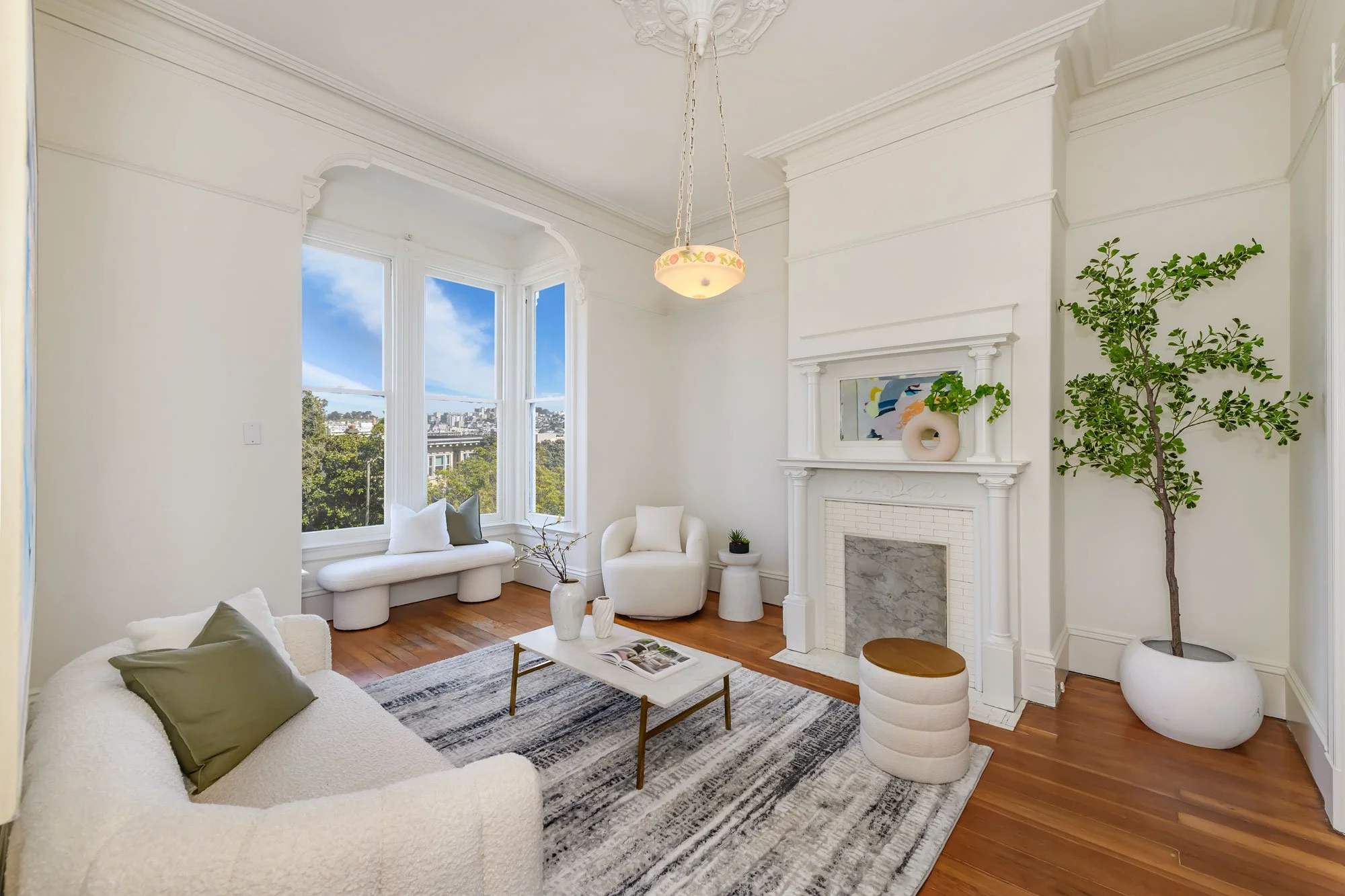 Bright living room with large bay window, white walls, fireplace, and modern furniture including a sofa with pillows, a white armchair, a coffee table, a small ottoman, and indoor plants, with hardwood floors and a view of a cityscape and blue sky se