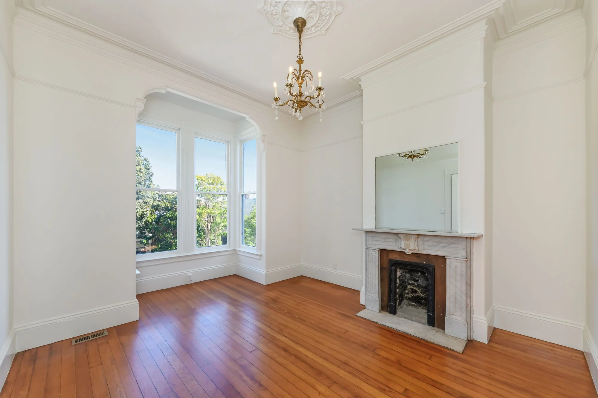 Empty room with white walls, hardwood floors, a fireplace with a mirror above it, large bay window, and a chandelier hanging from the ceiling.
