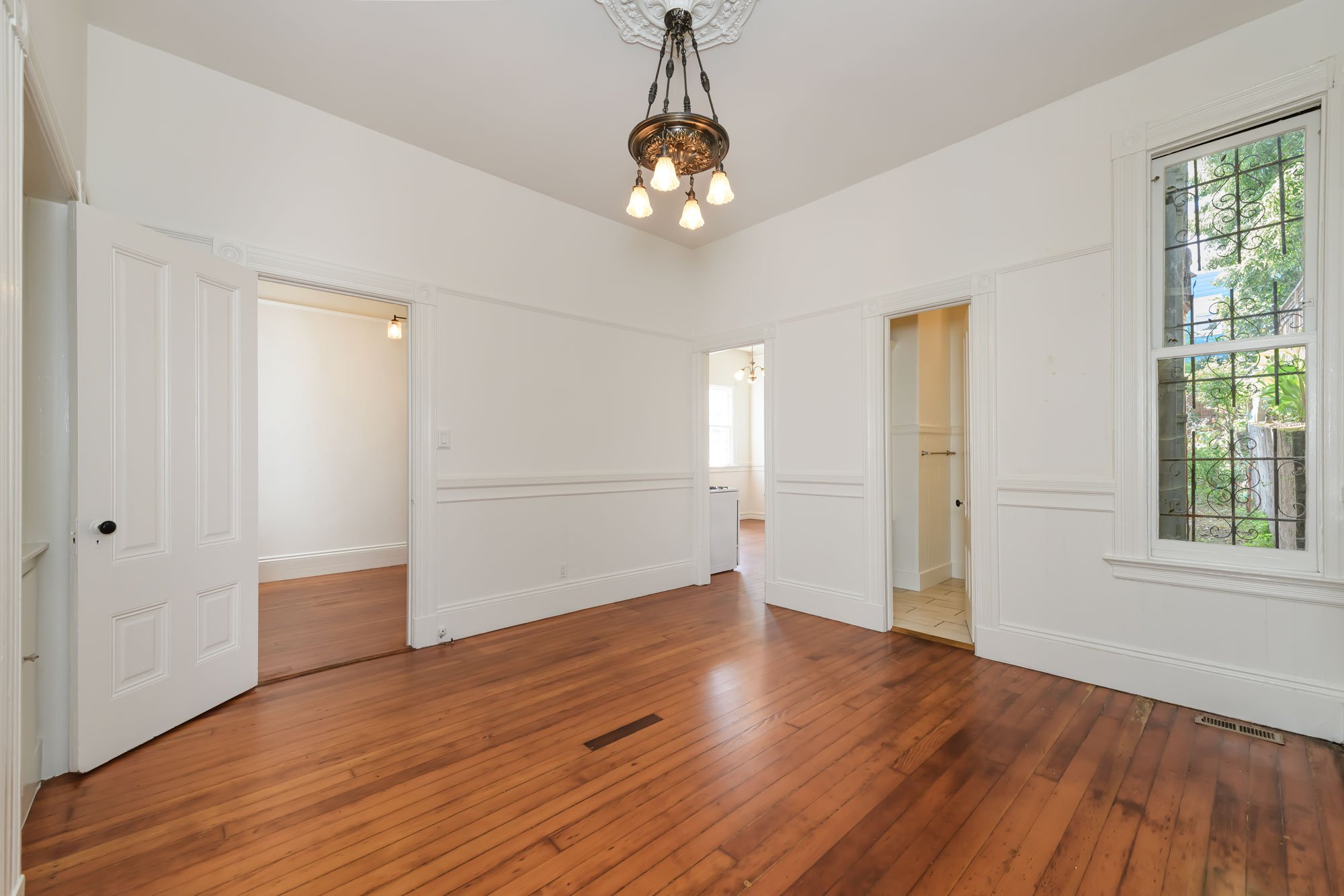 Empty living room with white walls, hardwood floors, a vintage chandelier, and large windows with decorative ironwork.