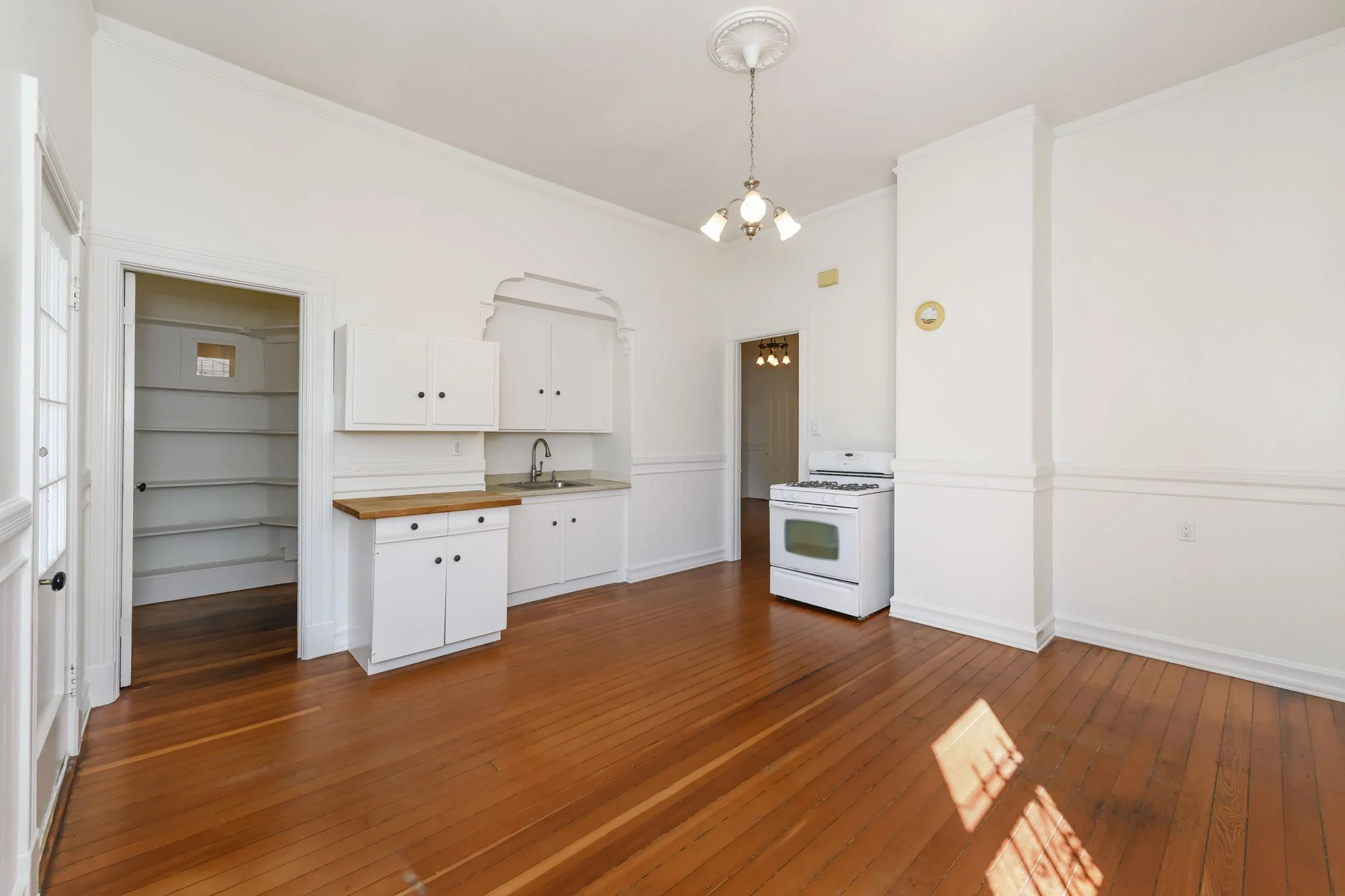 Empty kitchen with white cabinets, a wood countertop, a white stove, a stainless steel sink, and hardwood floors. There is a walk-in pantry with built-in shelves on the left.