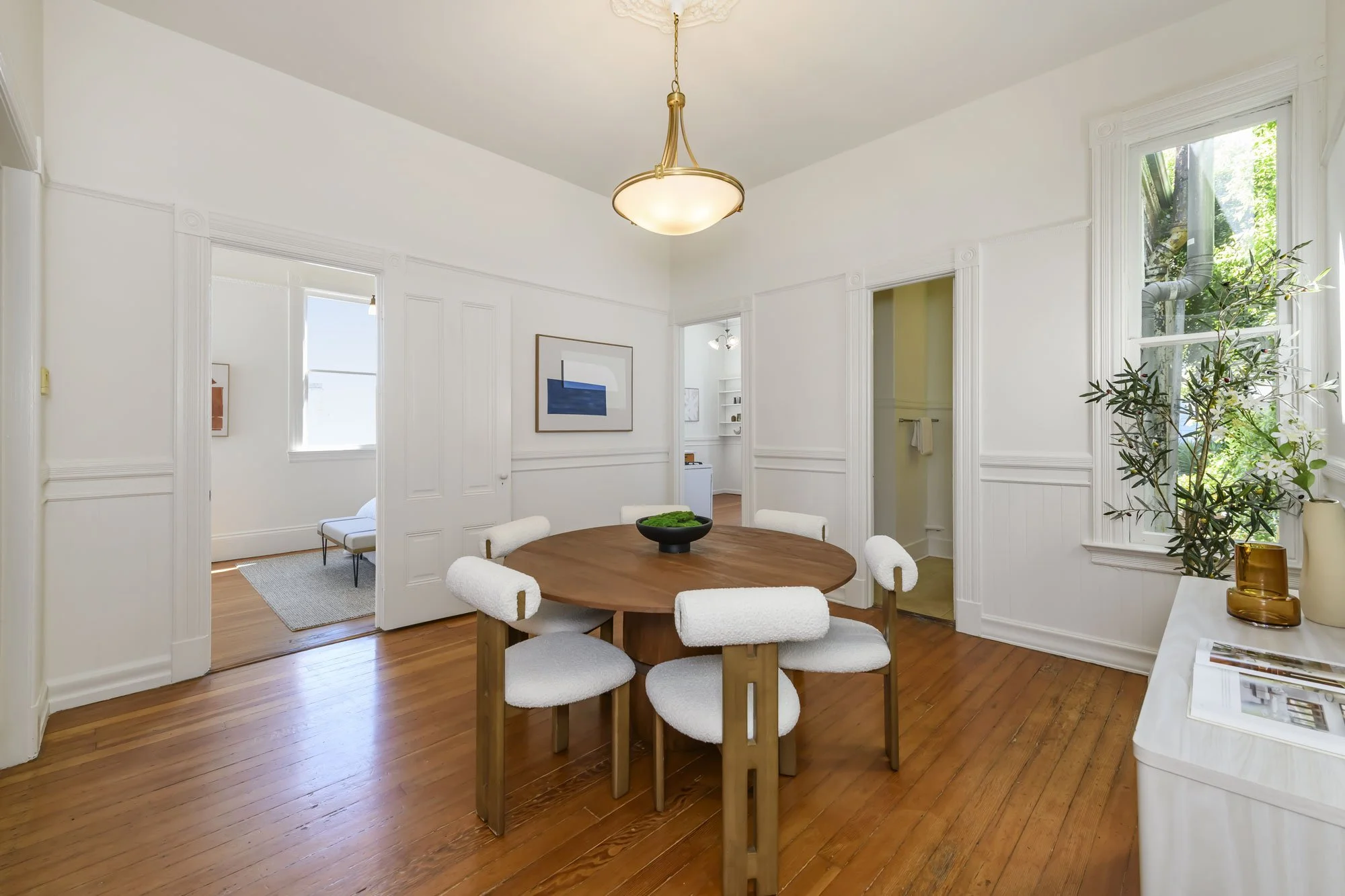 Dining room with white walls, a round wooden table with six white upholstered chairs, hardwood floors, a window with greenery outside, decorative plants and vases, and a doorway leading to another room with a chair and artwork.