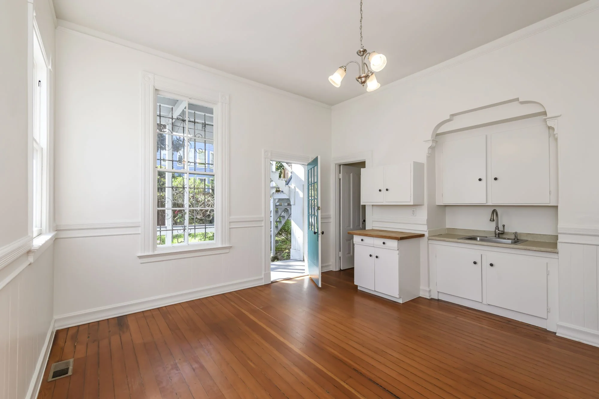 Empty kitchen or dining room with white walls, hardwood floors, a small white cabinet with a wooden top, a white sink, and a ceiling light fixture. An open door shows an outdoor staircase and garden.