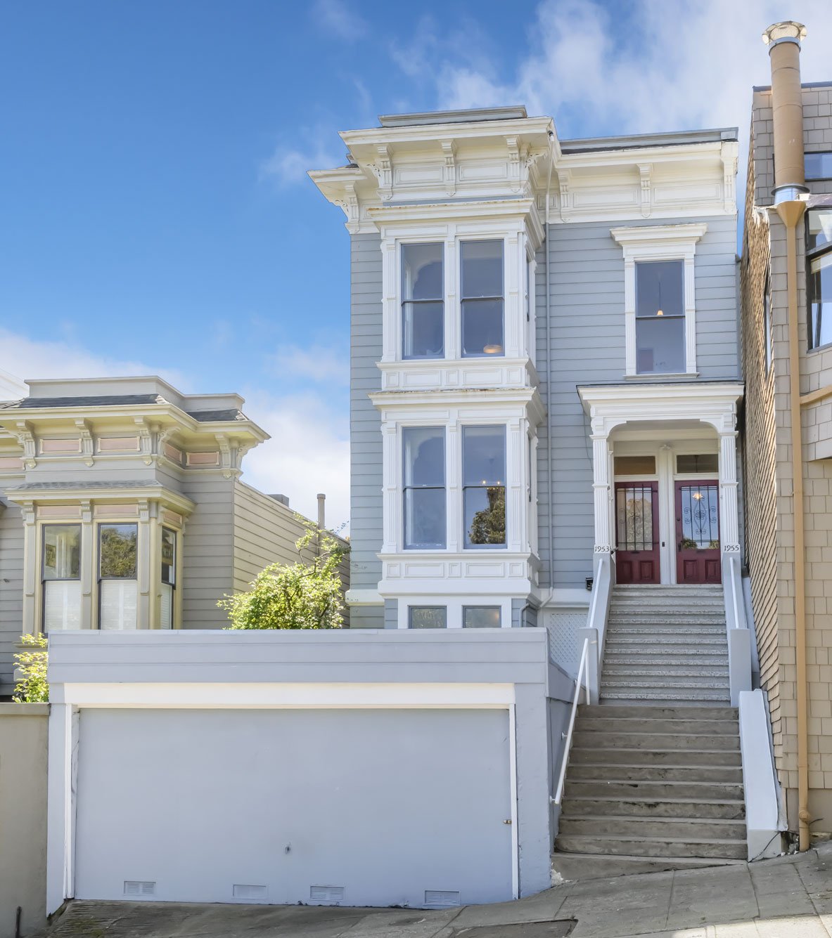Front view of a multi-story Victorian-style house with bay windows, painted light gray with white trim, red front doors, and stairs leading to the entrance, under a blue sky.