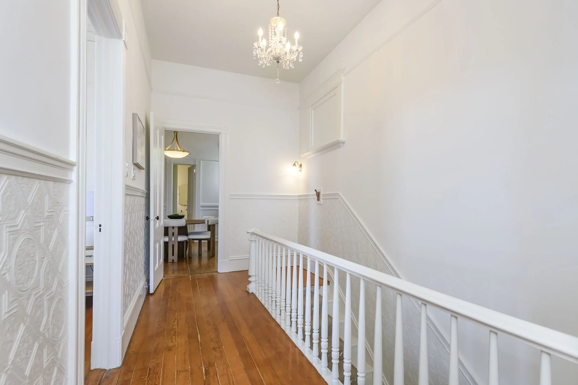 Bright hallway with white walls, wooden flooring, a chandelier hanging from the ceiling, and a staircase railing on the right. A doorway at the end of the hall leads to a dining area with a table and chairs.