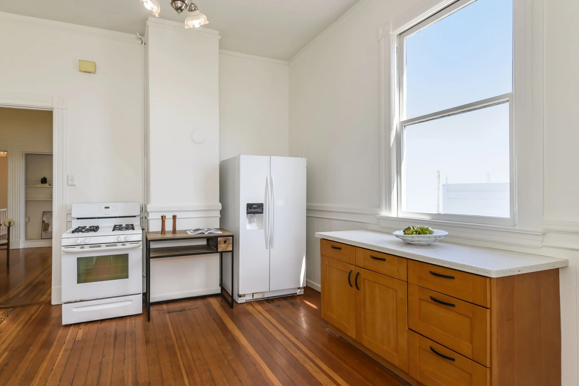A bright kitchen with white walls, hardwood floor, a white refrigerator, a white gas stove, a small black table with books on it, and a wooden cabinet with a white countertop near a large window.