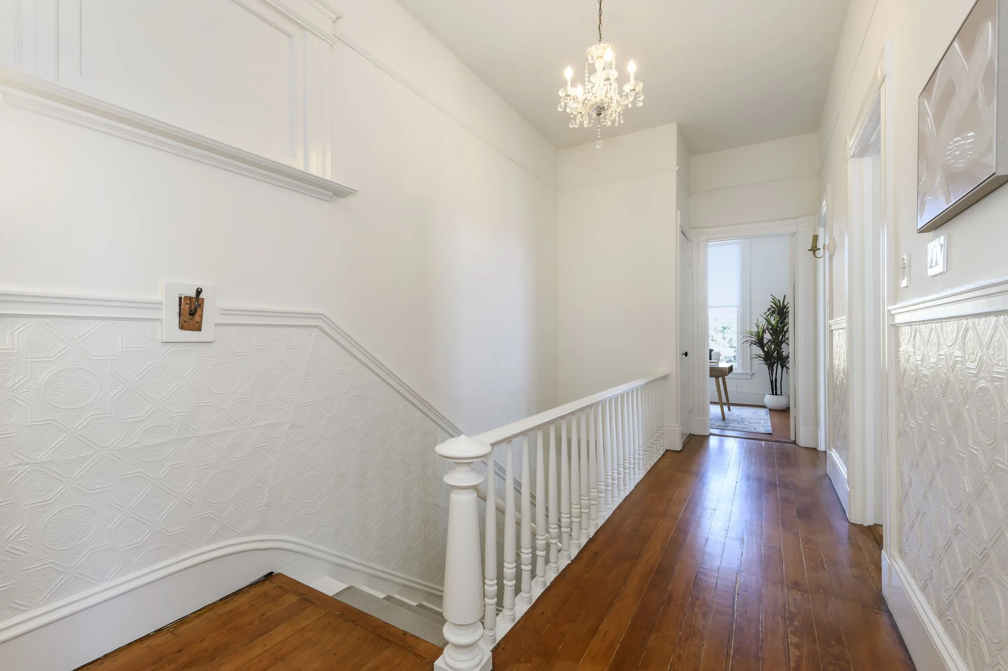 Bright hallway with white walls, wooden floor, a chandelier light fixture, and a stairway with white railing leading downstairs. At the end, an open doorway reveals a room with a large window, a potted plant, and a small table.