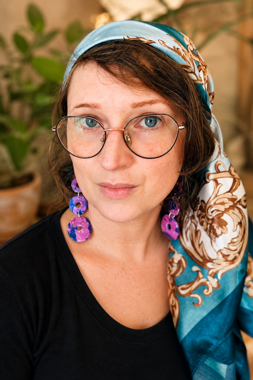 A woman with blue eyes and short brown hair, wearing large round glasses, purple earrings, and a patterned scarf on her head, sitting indoors with a plant in the background.