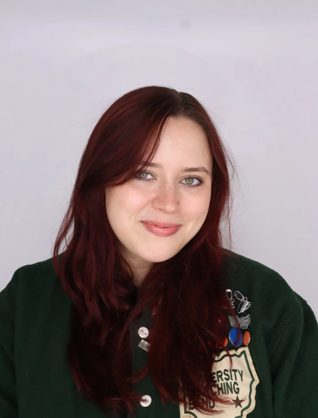 Smiling woman with red hair and light eyes, wearing a dark green sweater with pins and patches, standing against a light background.